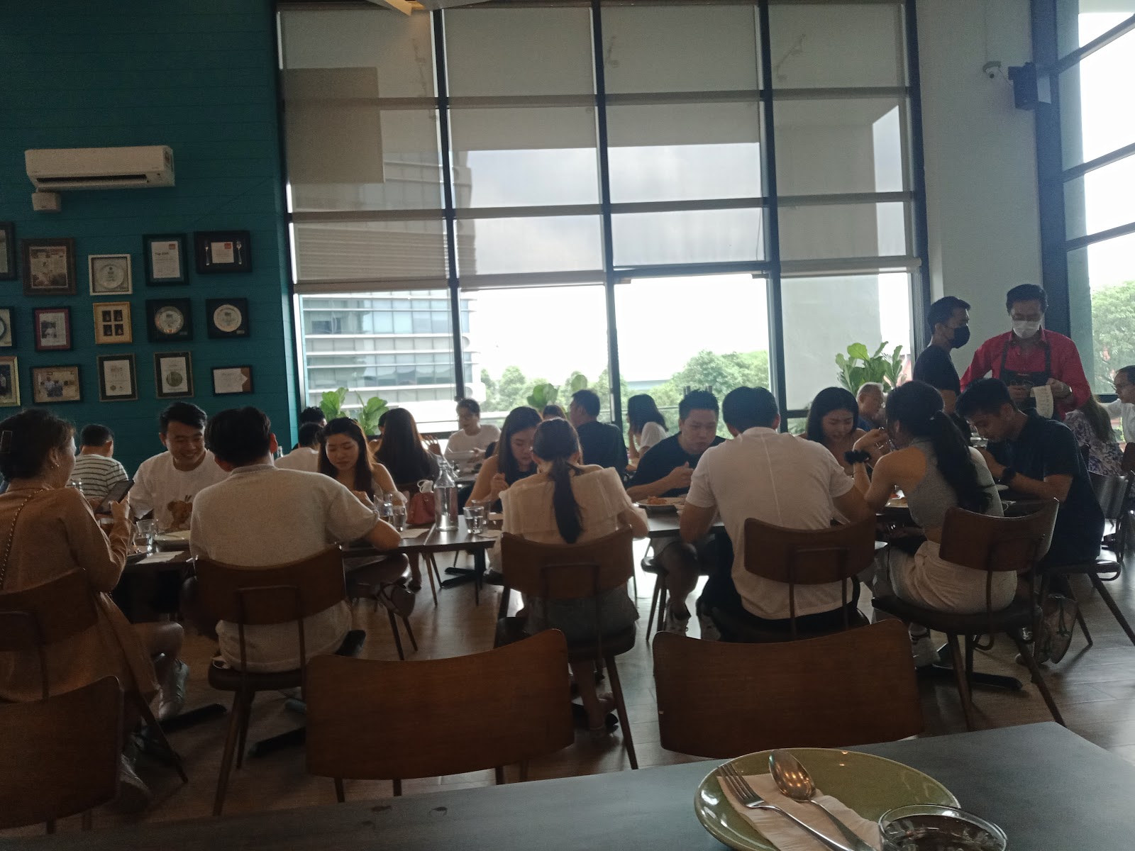 The Greens: A restaurant dining area filled with people seated at tables, near large glass windows overlooking greenery and buildings