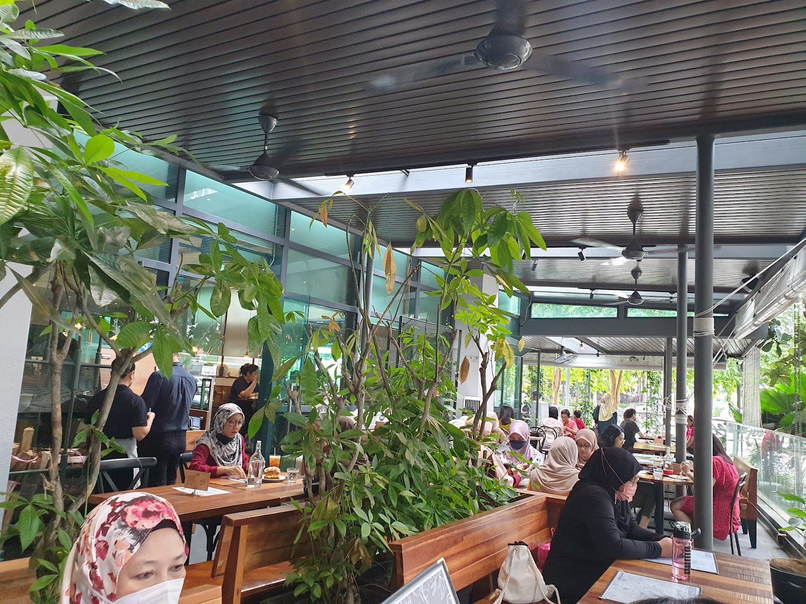 The Greens: An open-air café or restaurant seating area with wooden tables and benches, surrounded by lush plants, under a slatted ceiling with fans, with patrons dining