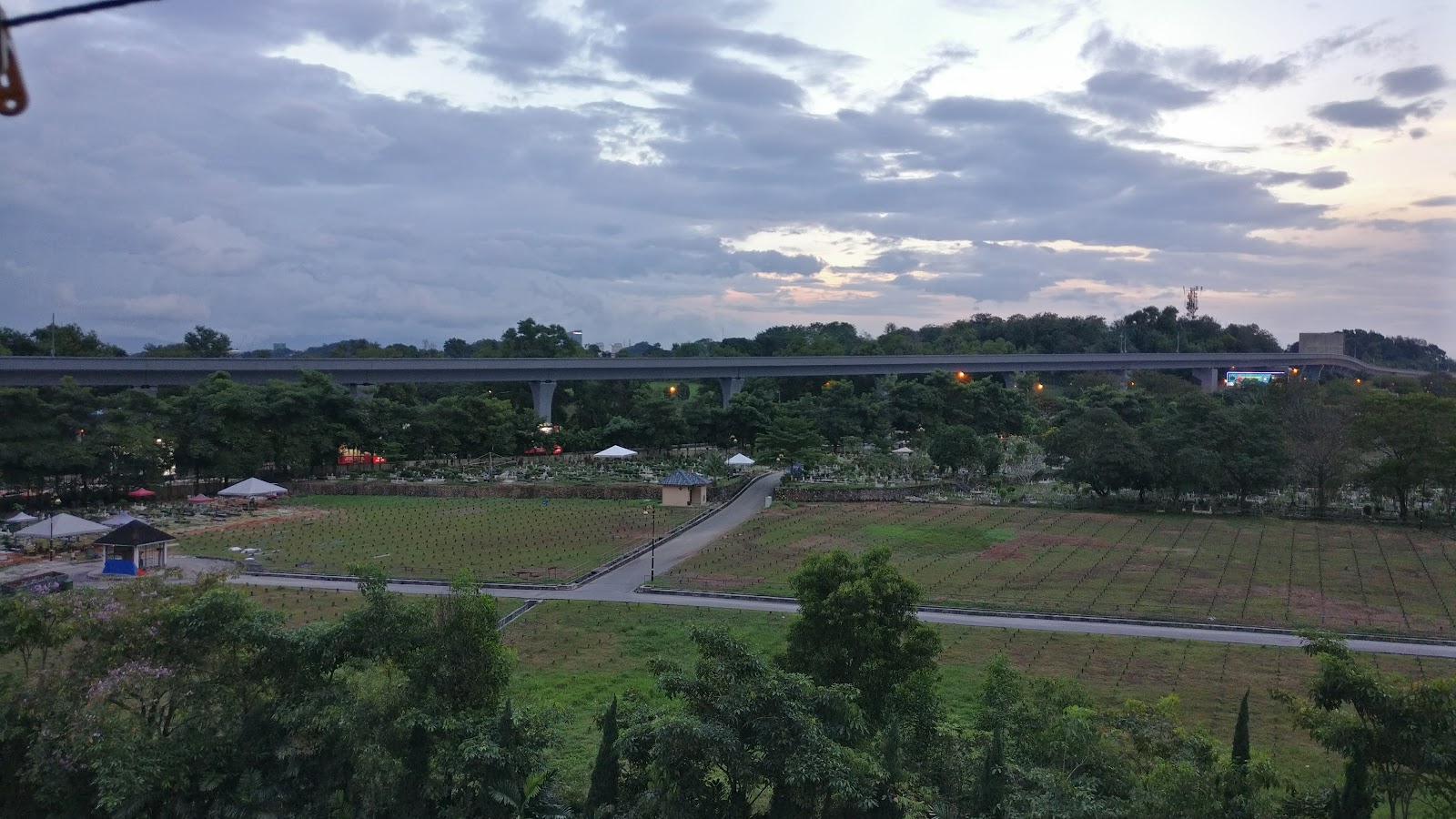 Desa Kiara: Elevated view overlooking a cemetery grounds with a highway bridge in the background under a cloudy sky