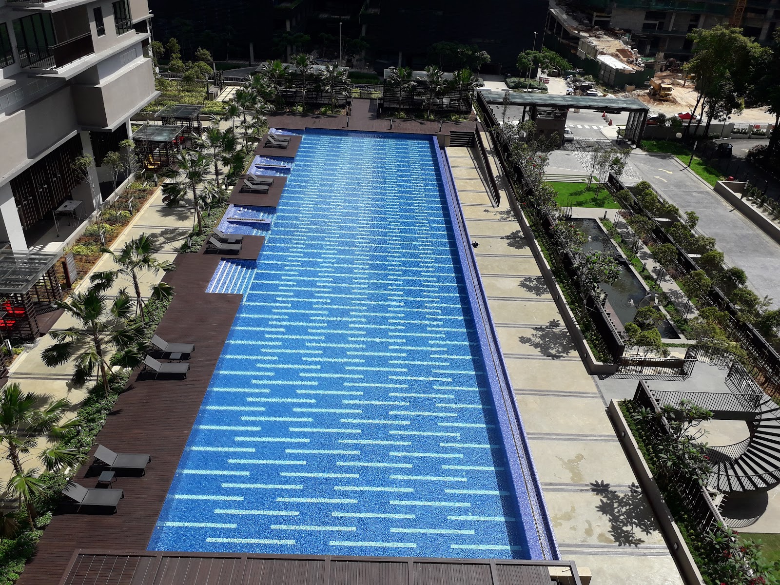 Residensi 22: Aerial view of a long lap swimming pool with lounge chairs on a wooden deck, surrounded by landscaped grounds of a residential development