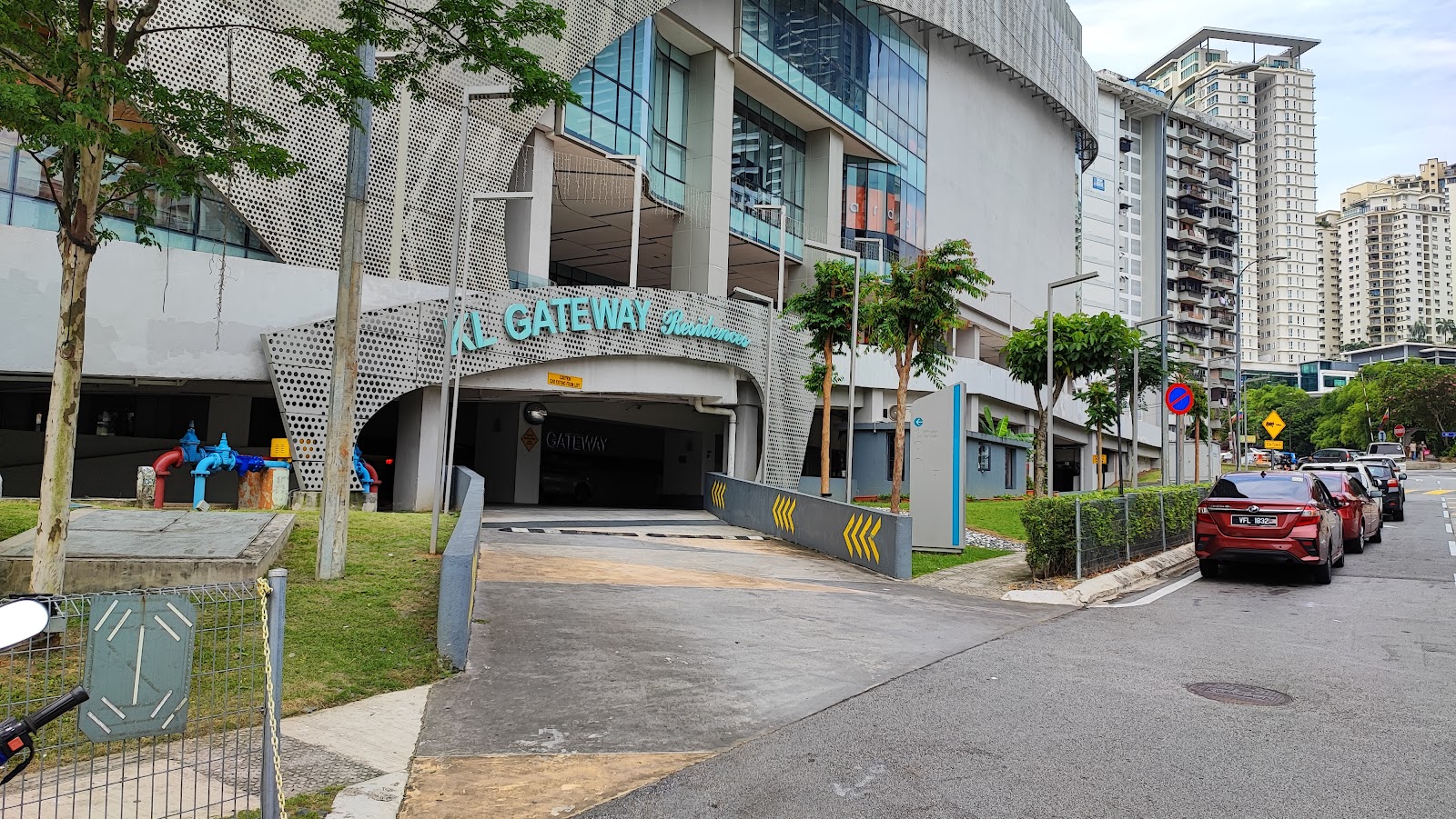KL Gateway Residences: Street view of the entrance driveway to a parking garage beneath KL Gateway Residences, with signage above the ramp and city towers in the background