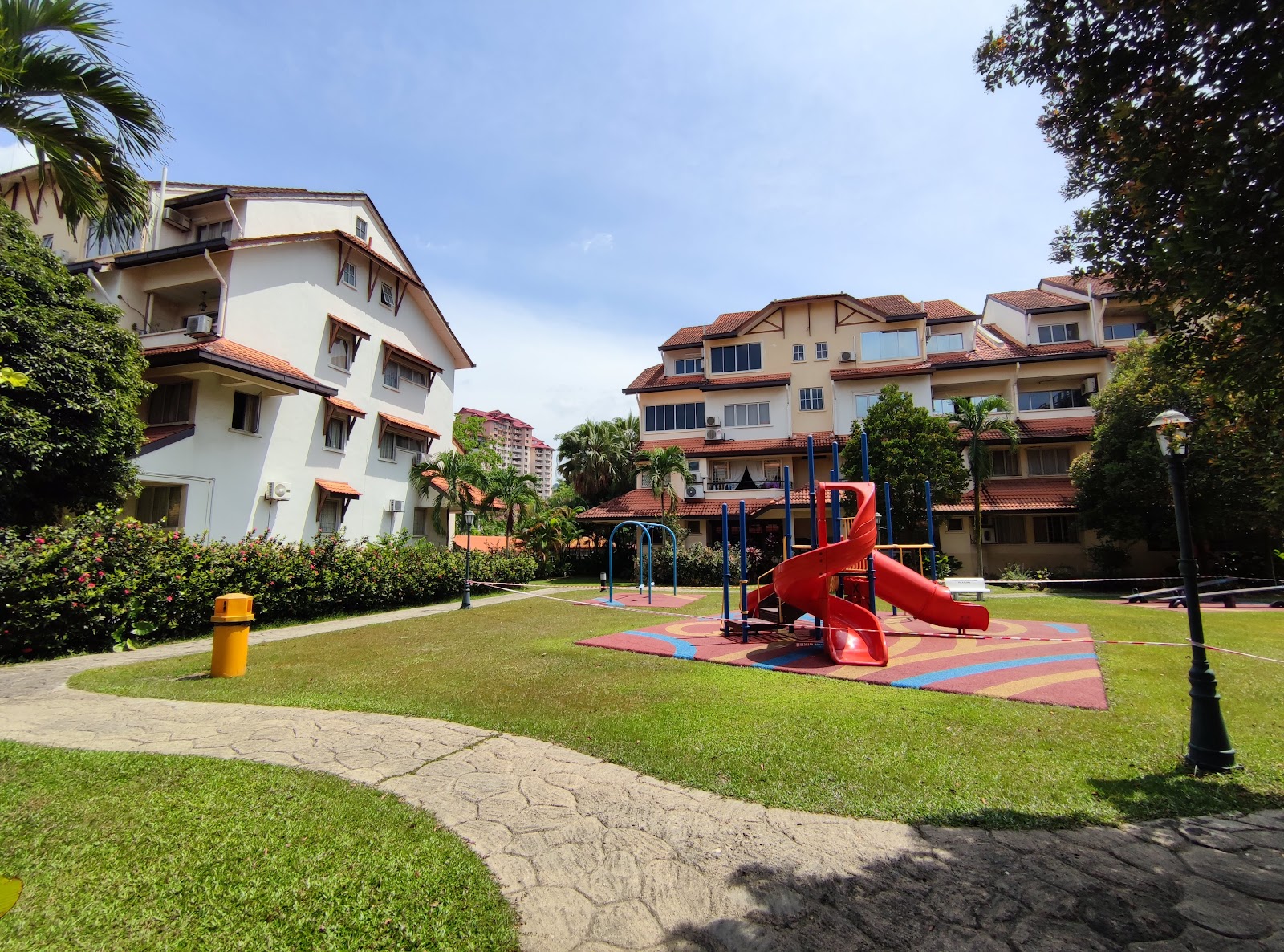 Villa Flora: A children's playground with a red slide and climbing equipment on rubberized flooring within a residential estate courtyard, surrounded by low-rise apartment buildings and greenery