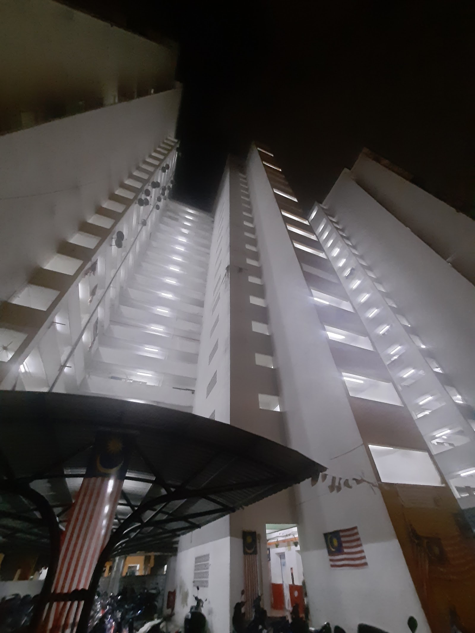PPR Pantai Ria: Night view looking straight up at tall apartment tower blocks with lit corridors and windows, taken from ground level near a covered entrance with Malaysian flags