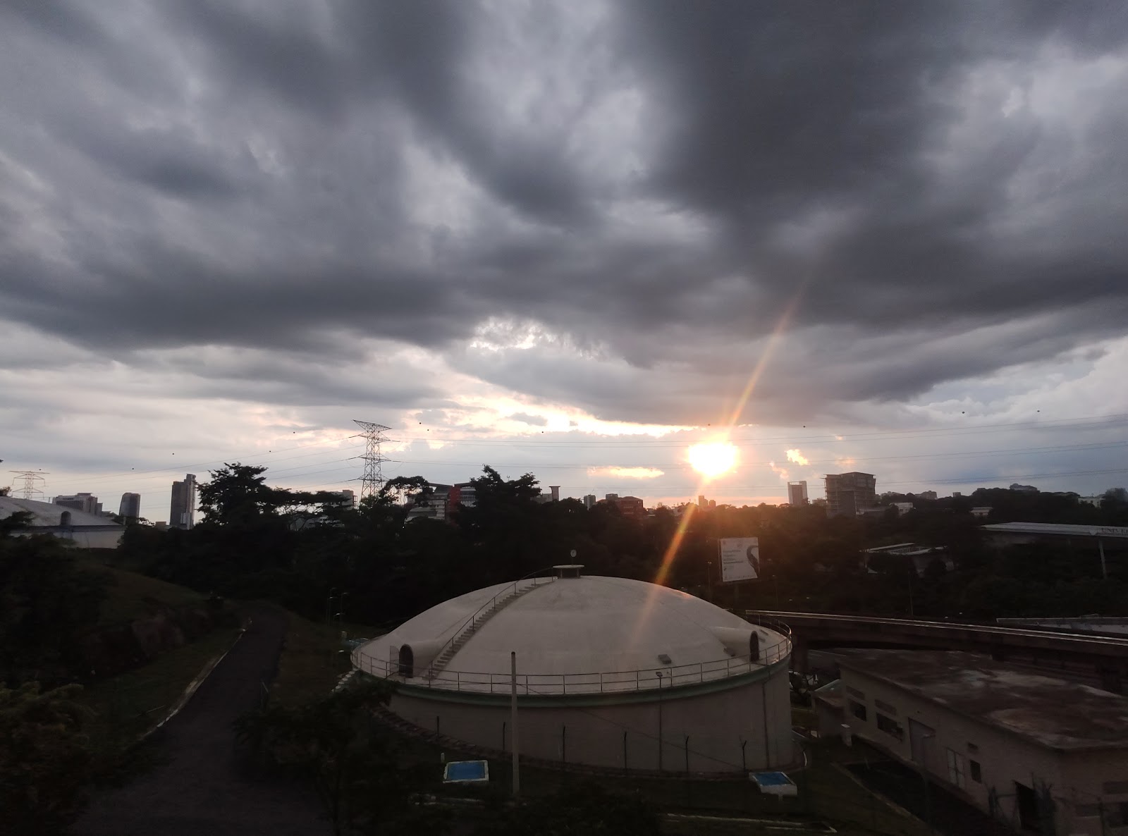 Vista Angkasa: Sunset sky over a cityscape with a rooftop water tank/dome structure in the foreground, silhouetted trees, transmission towers and buildings