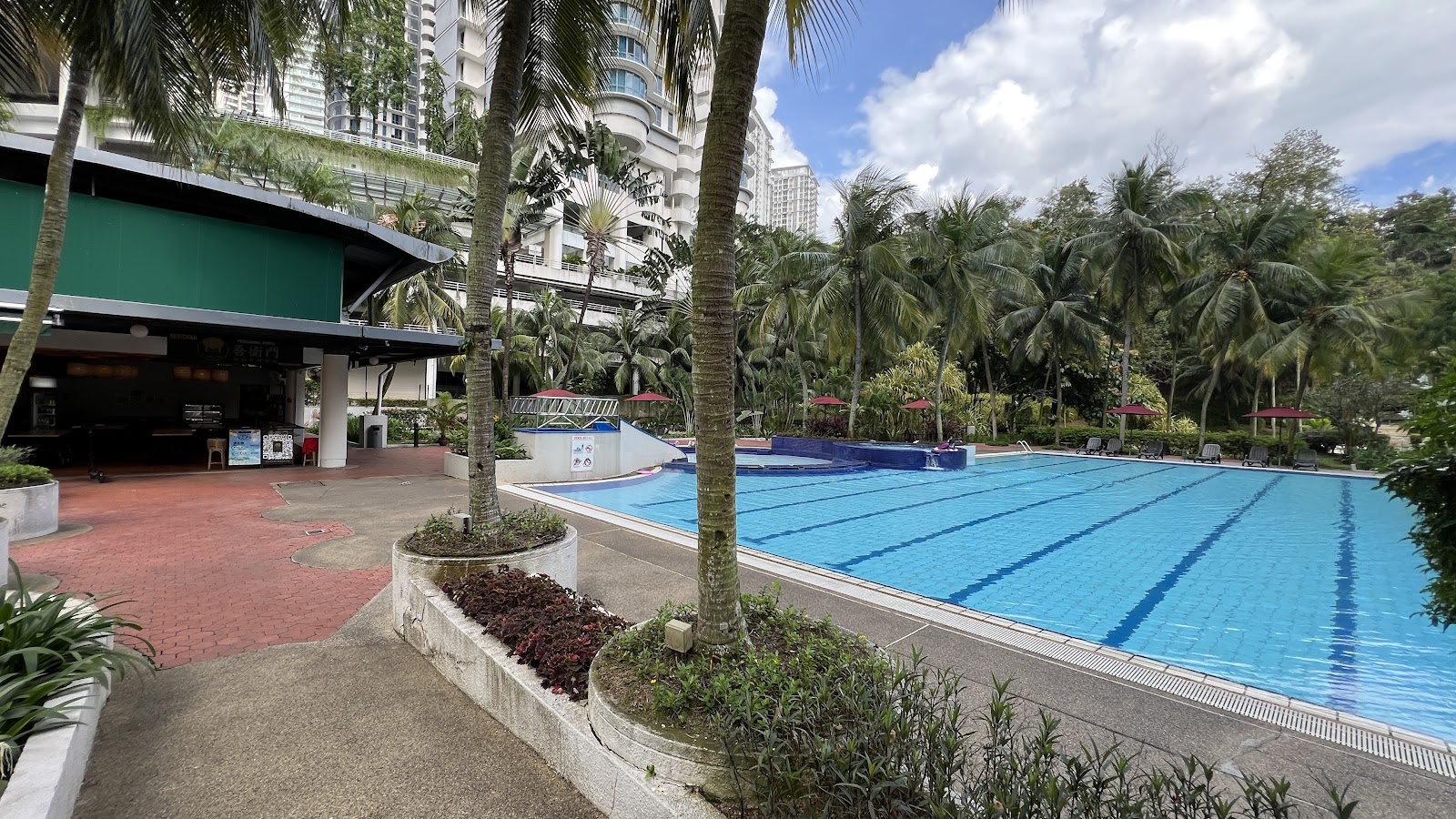 Mont Kiara Palma: Swimming pool area with lap lanes, surrounded by palm trees, lounge chairs and a covered poolside café, with high-rise towers in the background