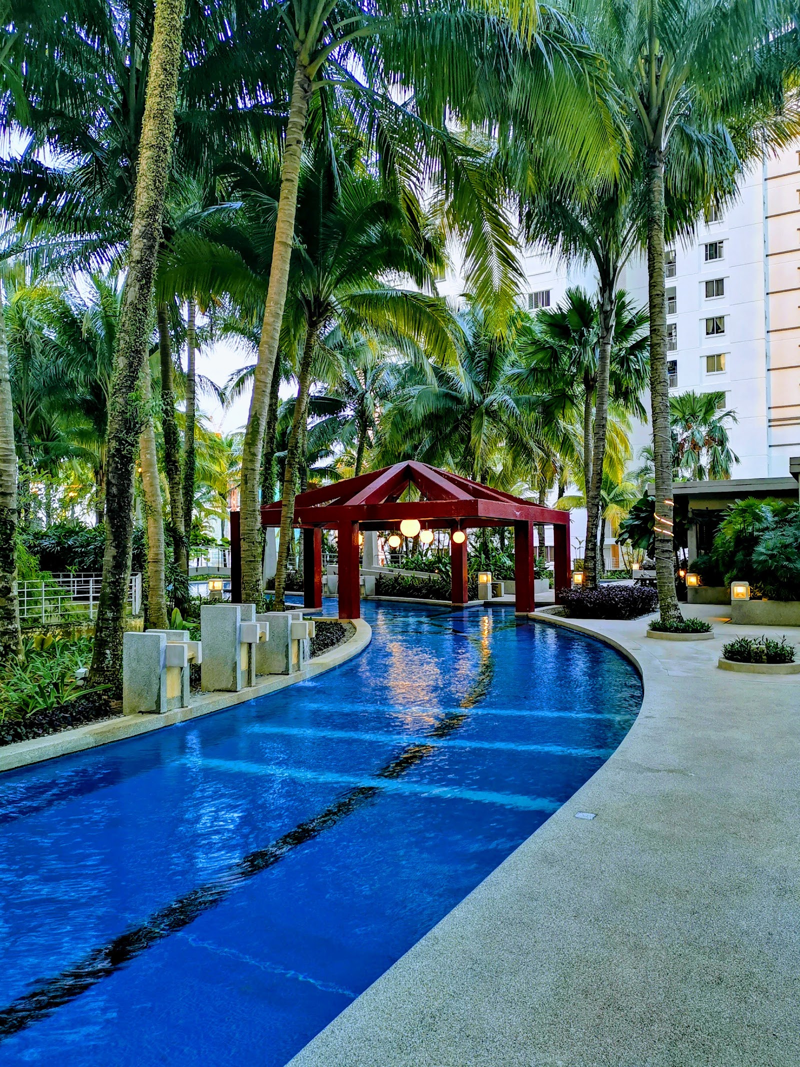 Kiaramas Ayuria: A curving resort-style lap pool lined with palm trees, with a red pergola structure over the pool and a high-rise building in the background