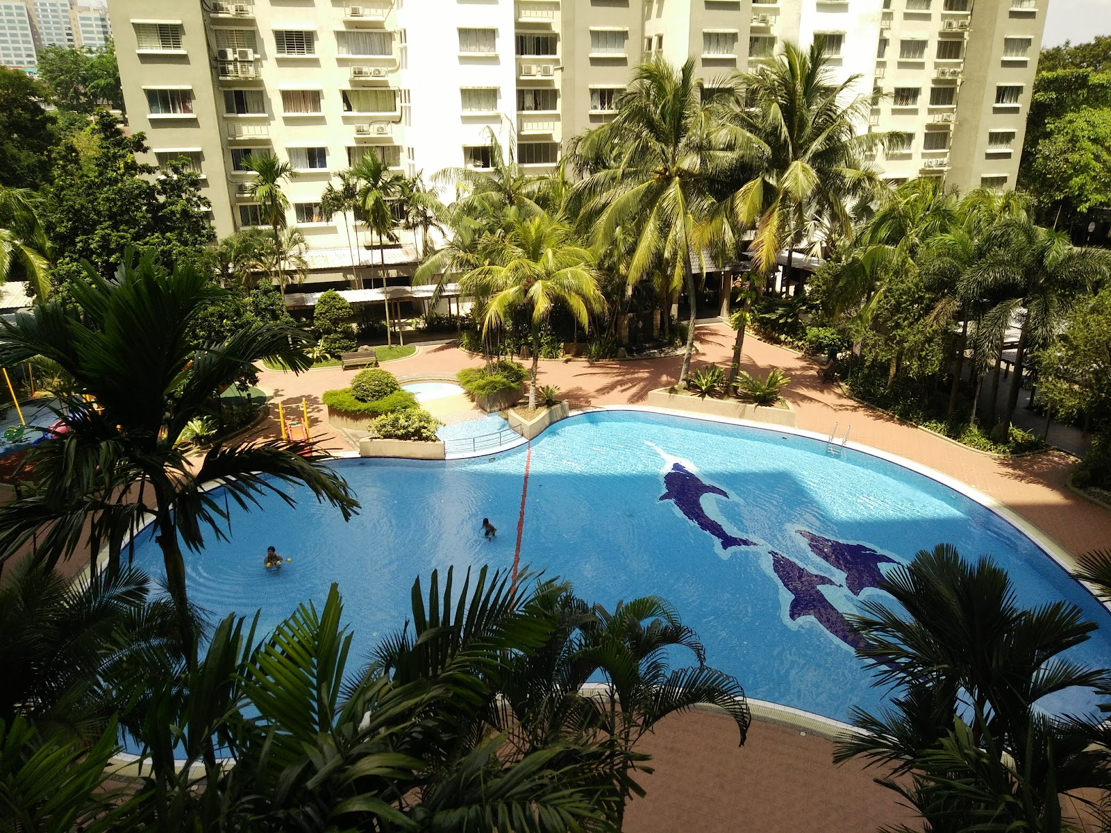 Puncak Damansara: Aerial view of a swimming pool with dolphin mosaic designs, surrounded by palm trees and landscaped grounds with a high-rise residential building in the background