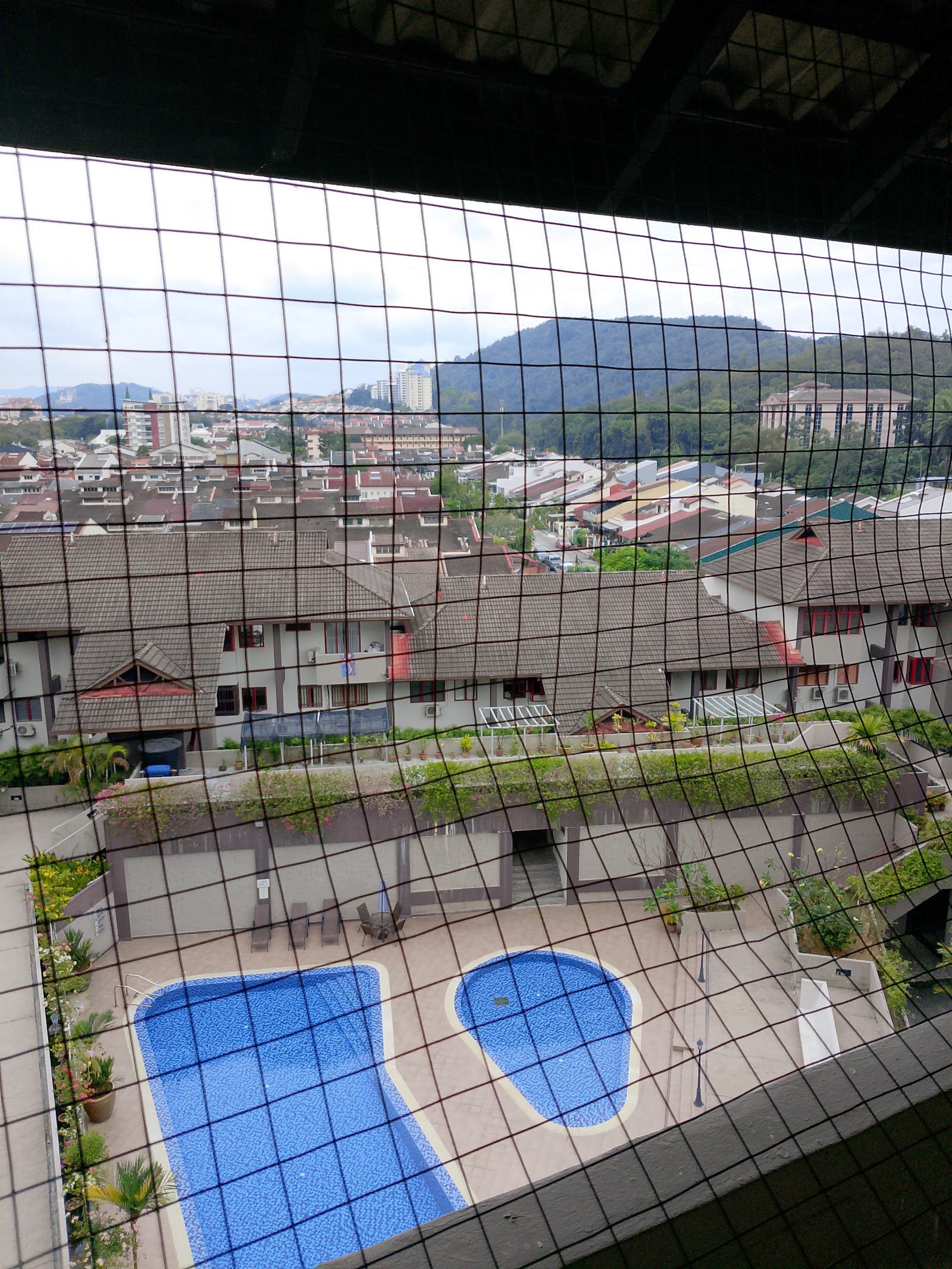Trellises: View through a netted balcony/window showing a rooftop pool area with two pools, loungers, and surrounding residential buildings and hills beyond