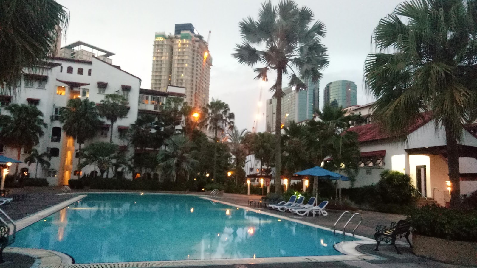 Pantai Hillpark 2: Outdoor swimming pool with loungers and umbrellas at dusk, surrounded by palm trees, the property's buildings, and city skyscrapers in the background