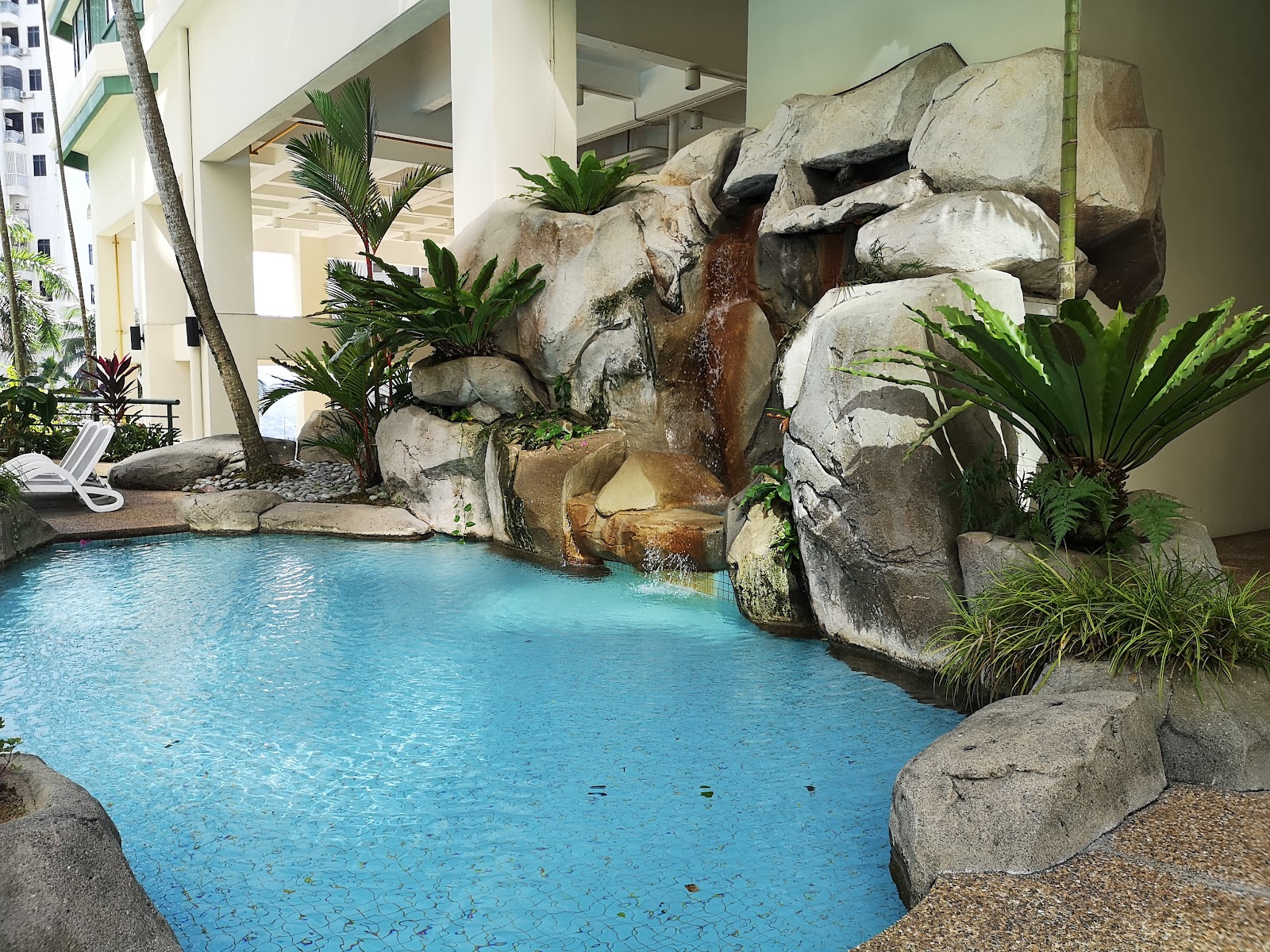 Araville: A swimming pool with a rock waterfall feature, tropical landscaping, and a lounge chair beside a building