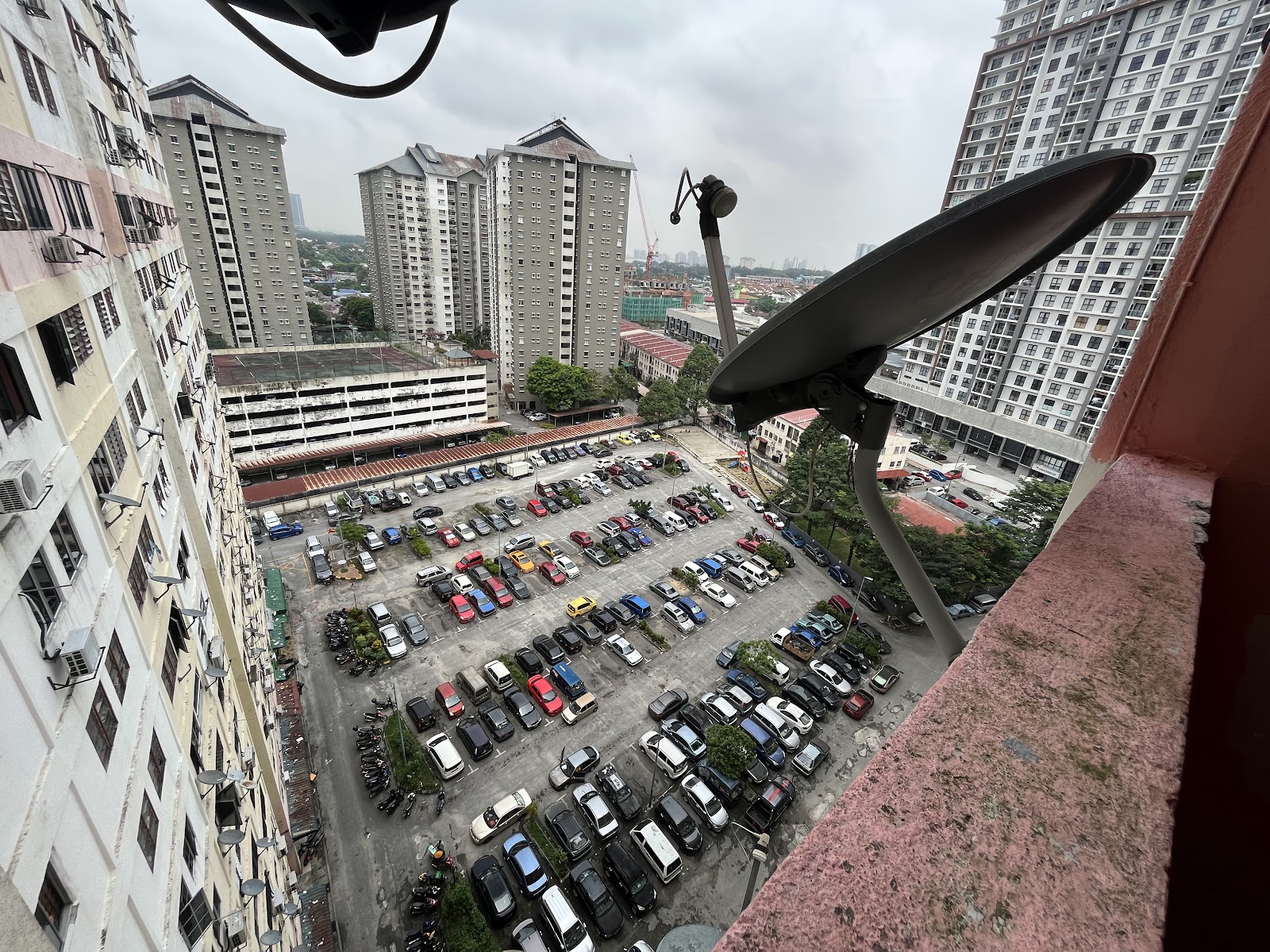 Flat Kayu Ara: View from a high-rise balcony overlooking a large parking lot surrounded by residential tower blocks, with a satellite dish mounted in the foreground
