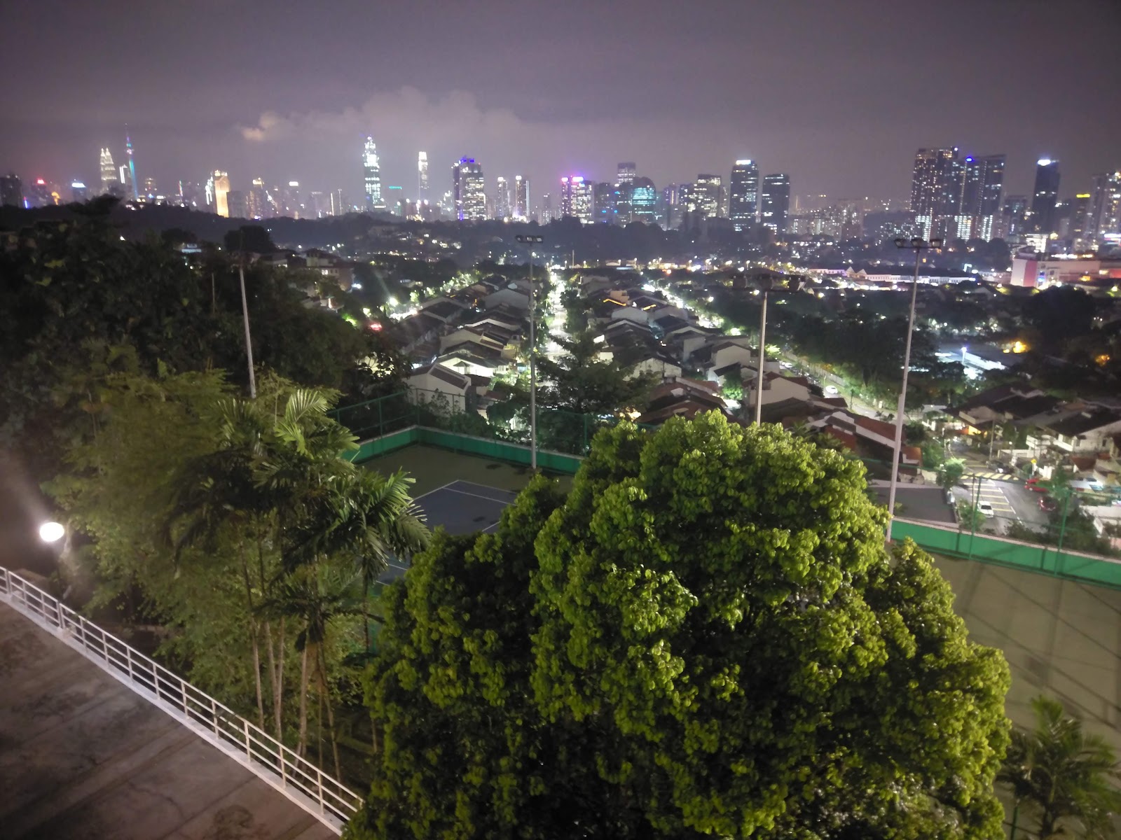 Sri Wangsaria: Night-time cityscape view from a high-rise balcony, overlooking trees, a rooftop court, residential houses, and distant skyscrapers