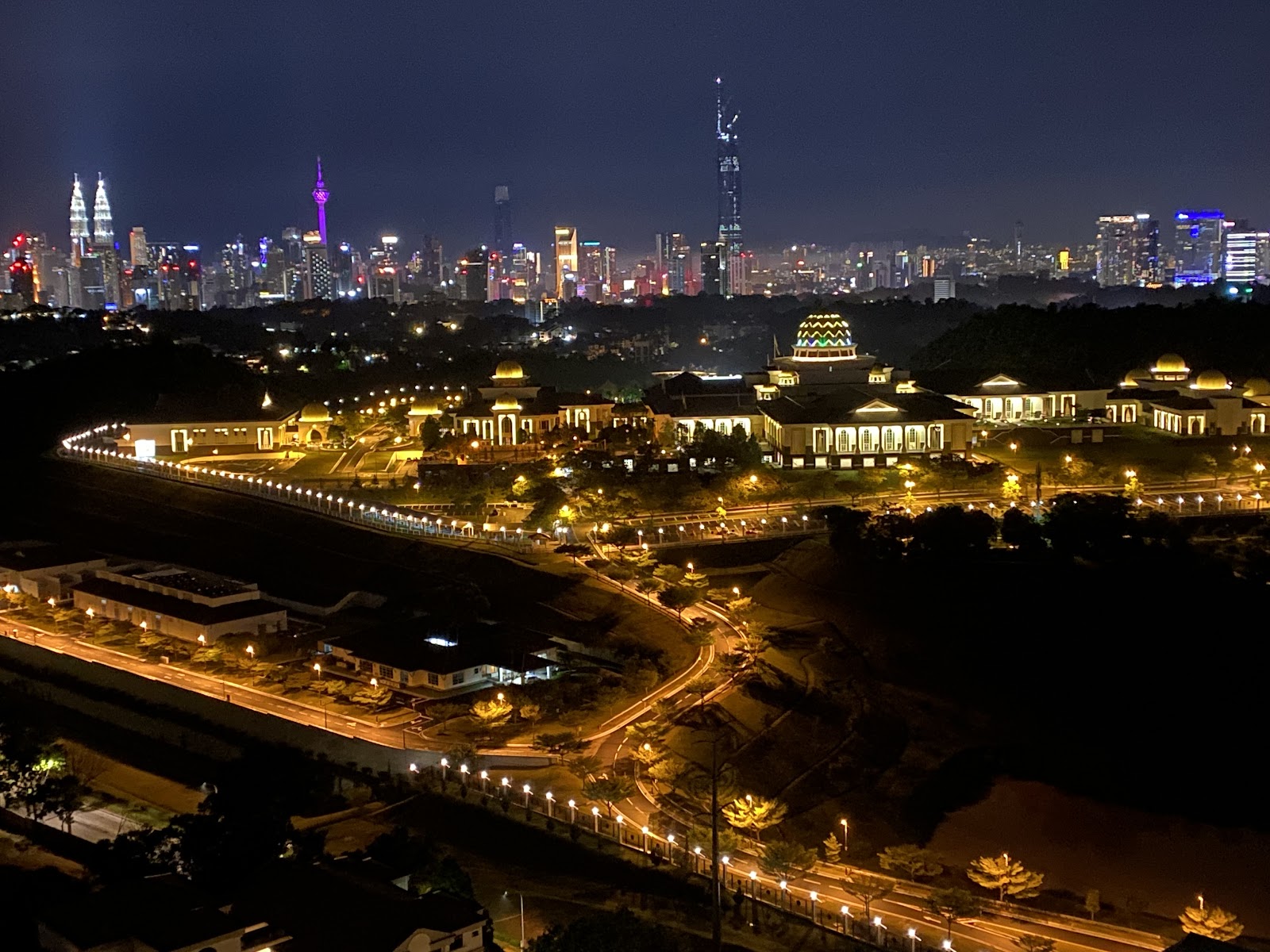 Windsor Tower: Nighttime aerial view from a high-rise showing a domed palace complex illuminated below and the Kuala Lumpur city skyline with Petronas Towers and KL Tower in the distance