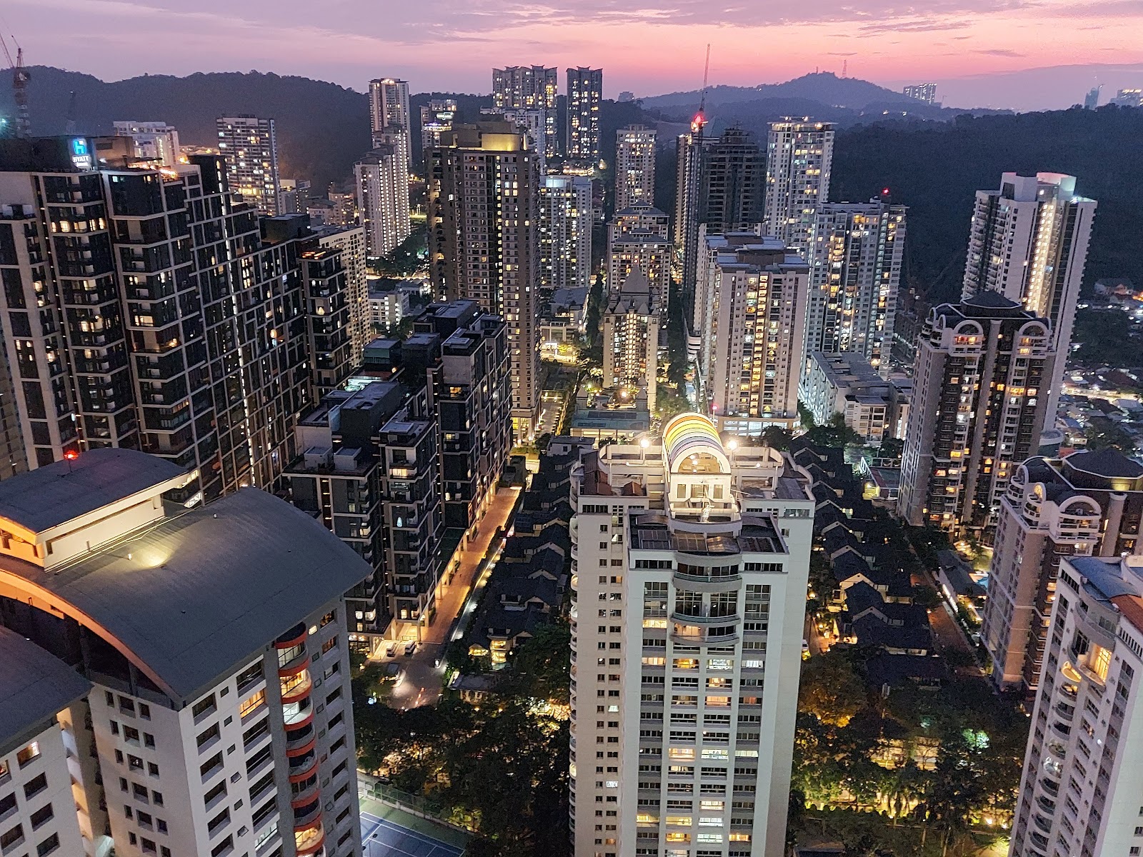Mont Kiara Pelangi: Aerial night view of a dense cityscape of illuminated high-rise residential towers with hills in the background, taken from a high floor of a development at dusk