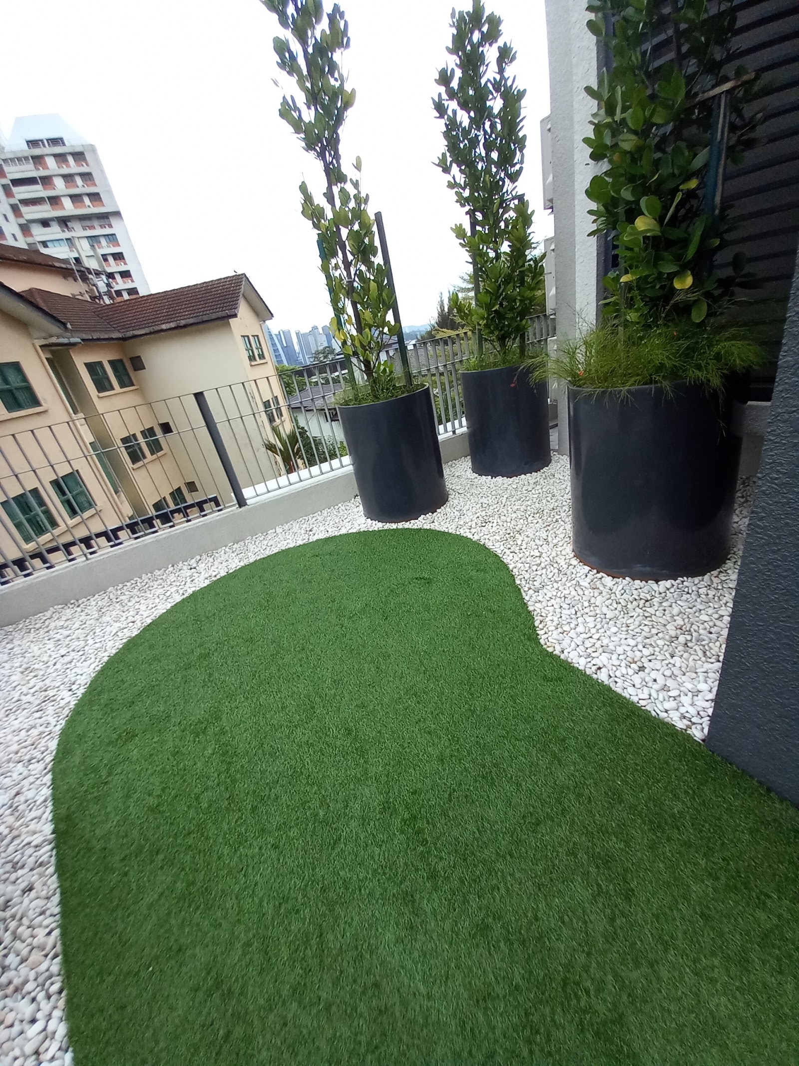 6 Kapas: A rooftop garden terrace with artificial grass, white pebbles, and large black planters with topiary trees, overlooking neighboring buildings