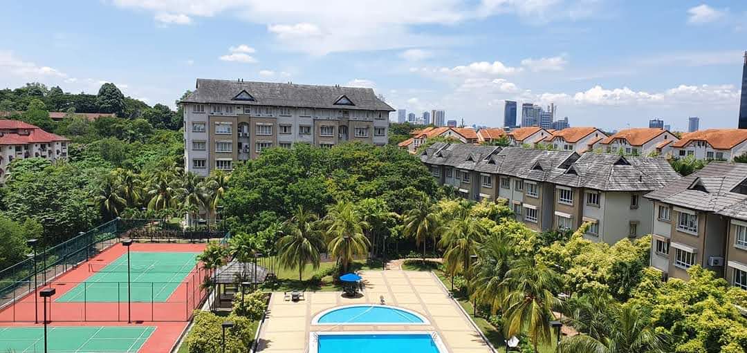 Sri TTDI: Aerial view of a residential development showing tennis courts, a swimming pool, and surrounding low-rise buildings amid greenery with a city skyline in the background