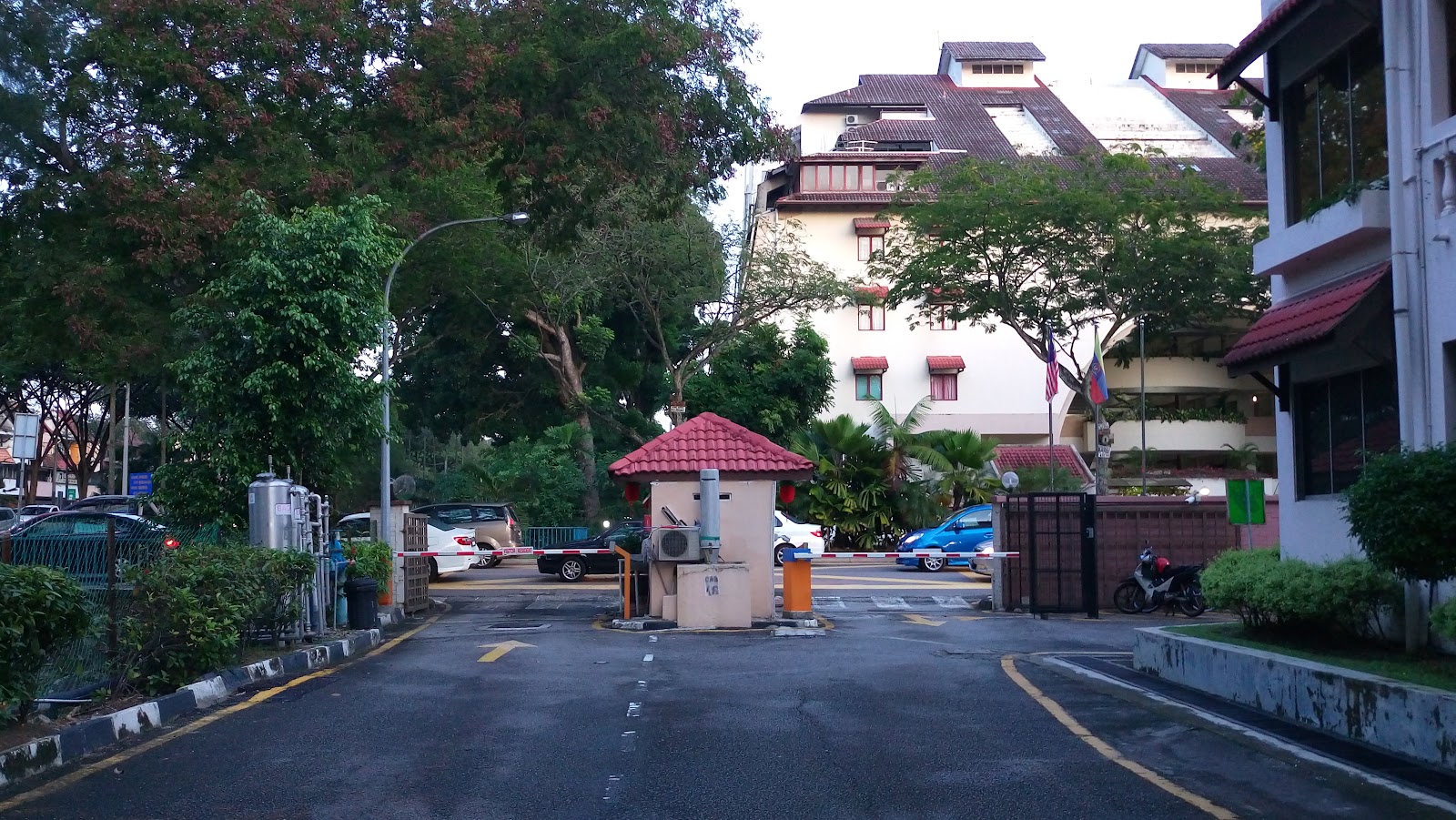 Kiara Green Townhouses: A street view of a residential development entrance with a guardhouse, barrier gate, parked cars, trees, and a multi-story building with red roofing in the background.