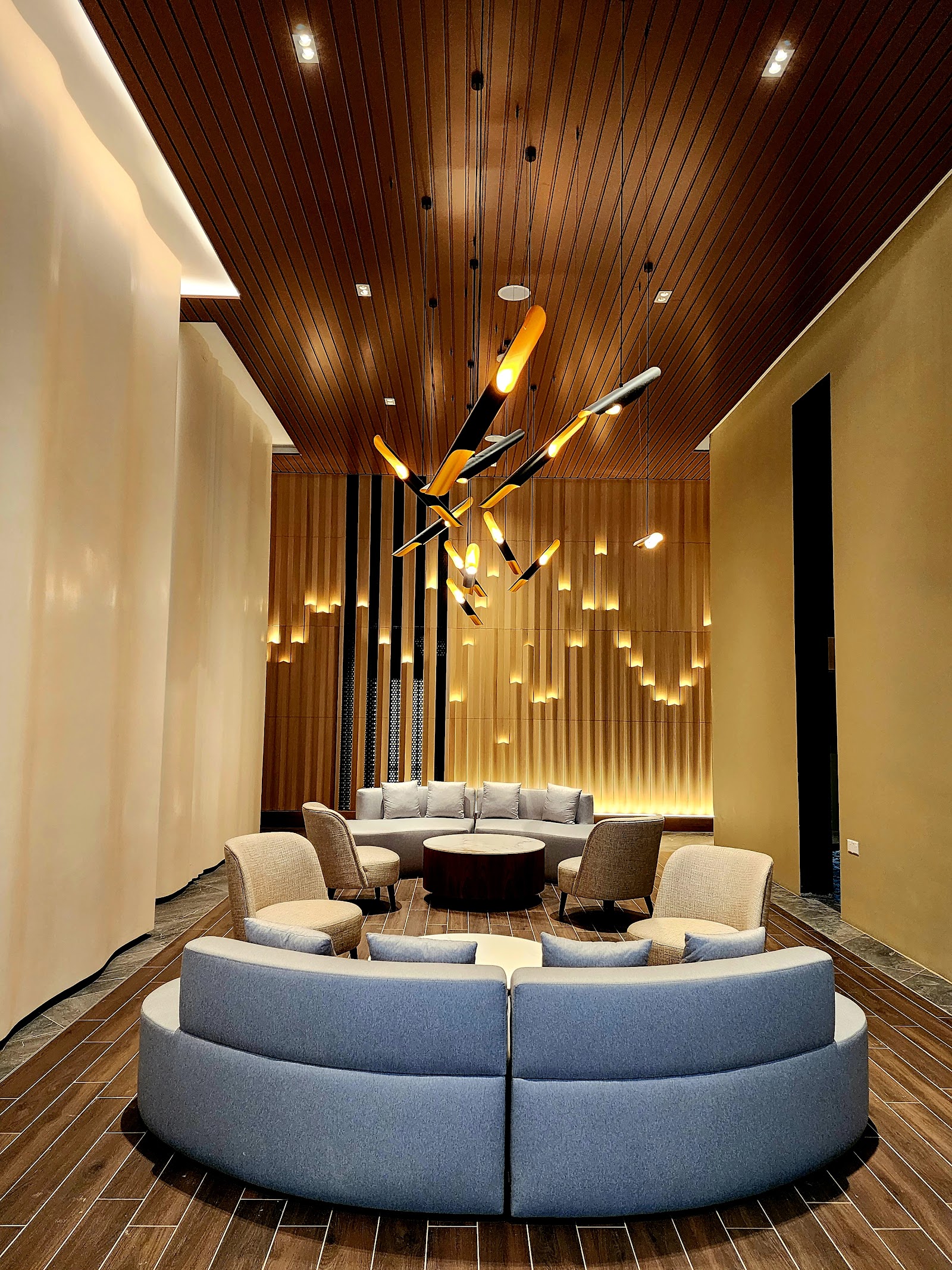 Allevia: A lobby or lounge waiting area with circular blue and beige sofas, a round coffee table, decorative hanging light fixtures, and wood-paneled ceiling and wall accents