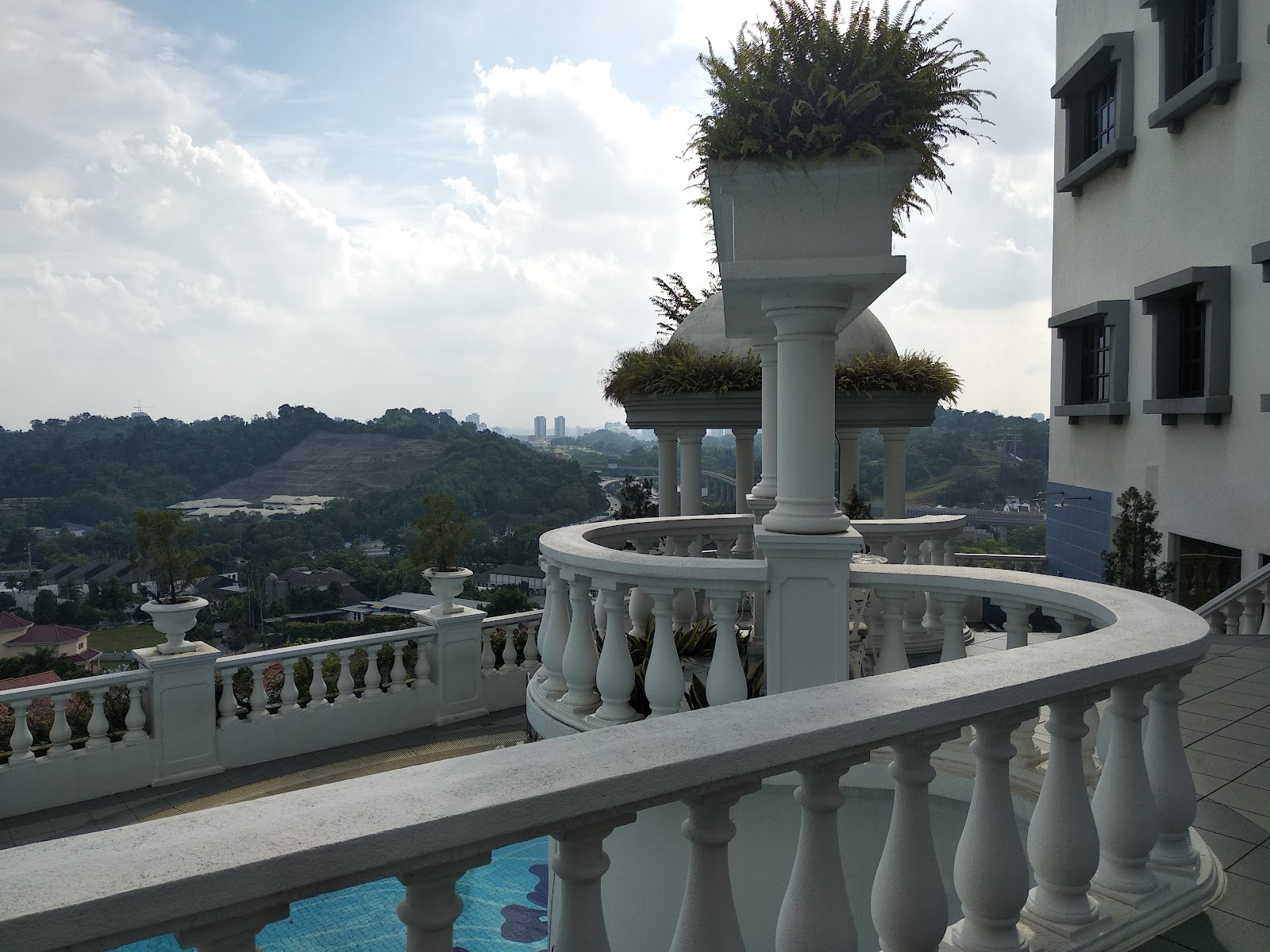 Contessa: A curved white balustrade balcony with ornate pillars and planters overlooking a pool, hillside, trees, and distant city skyline