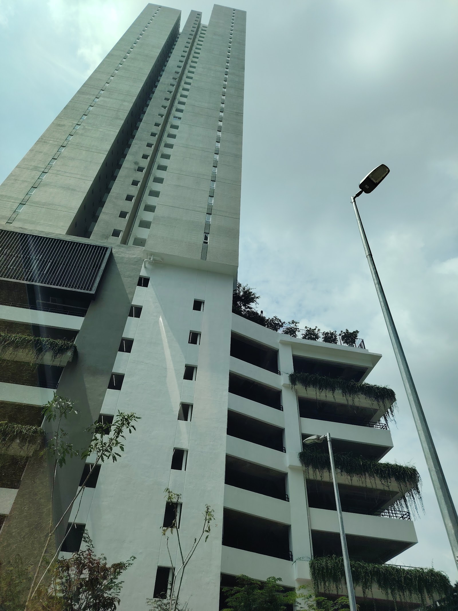 Residensi Dutamas Dahlia: Upward view of a tall residential tower with a lower balcony wing featuring greenery, seen from ground level against a cloudy sky, with a streetlamp in foreground