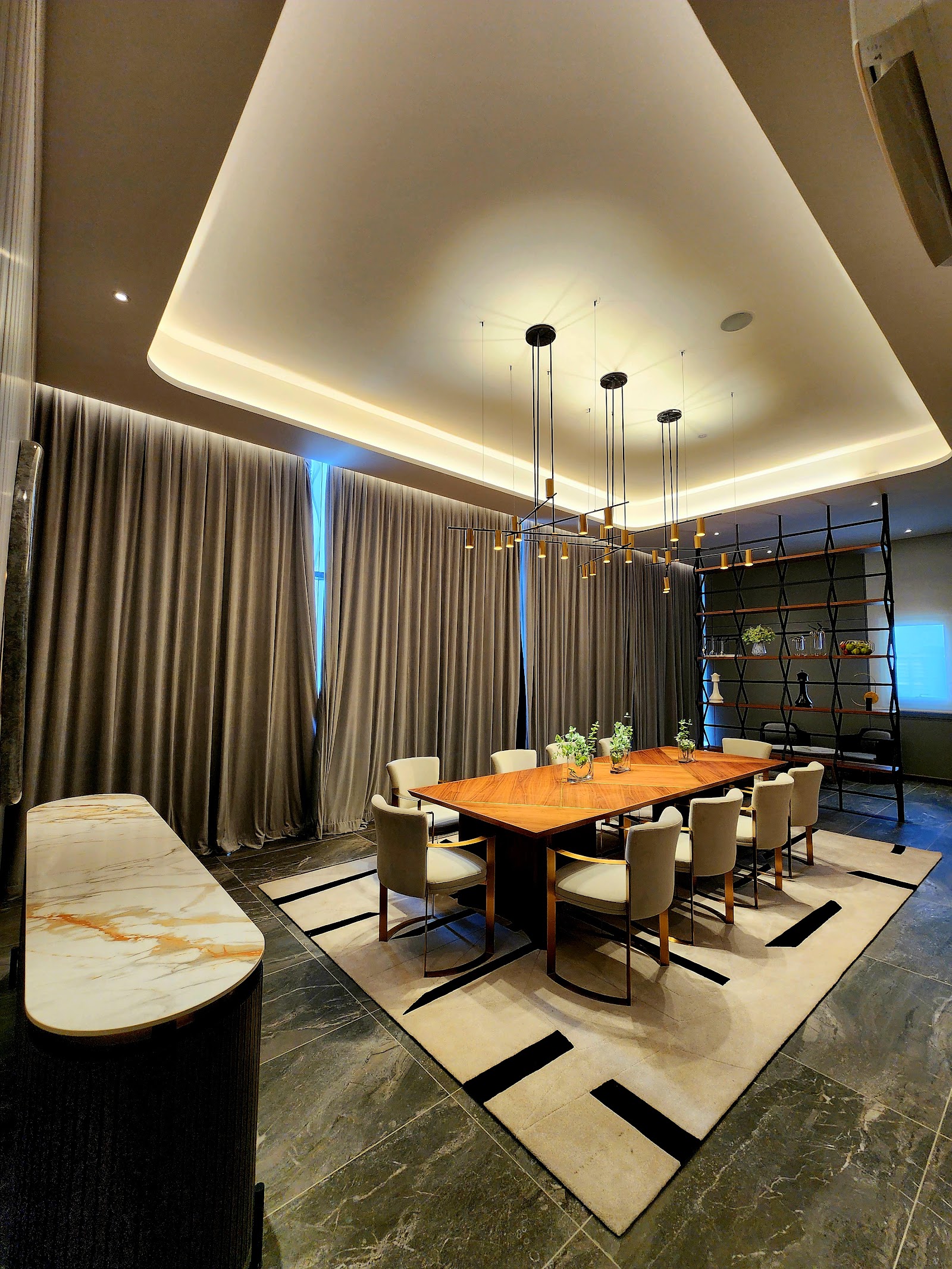 Allevia: An elegant dining function room with a long wooden table, upholstered chairs, marble console, decorative shelving, and a suspended pendant light fixture under a recessed ceiling
