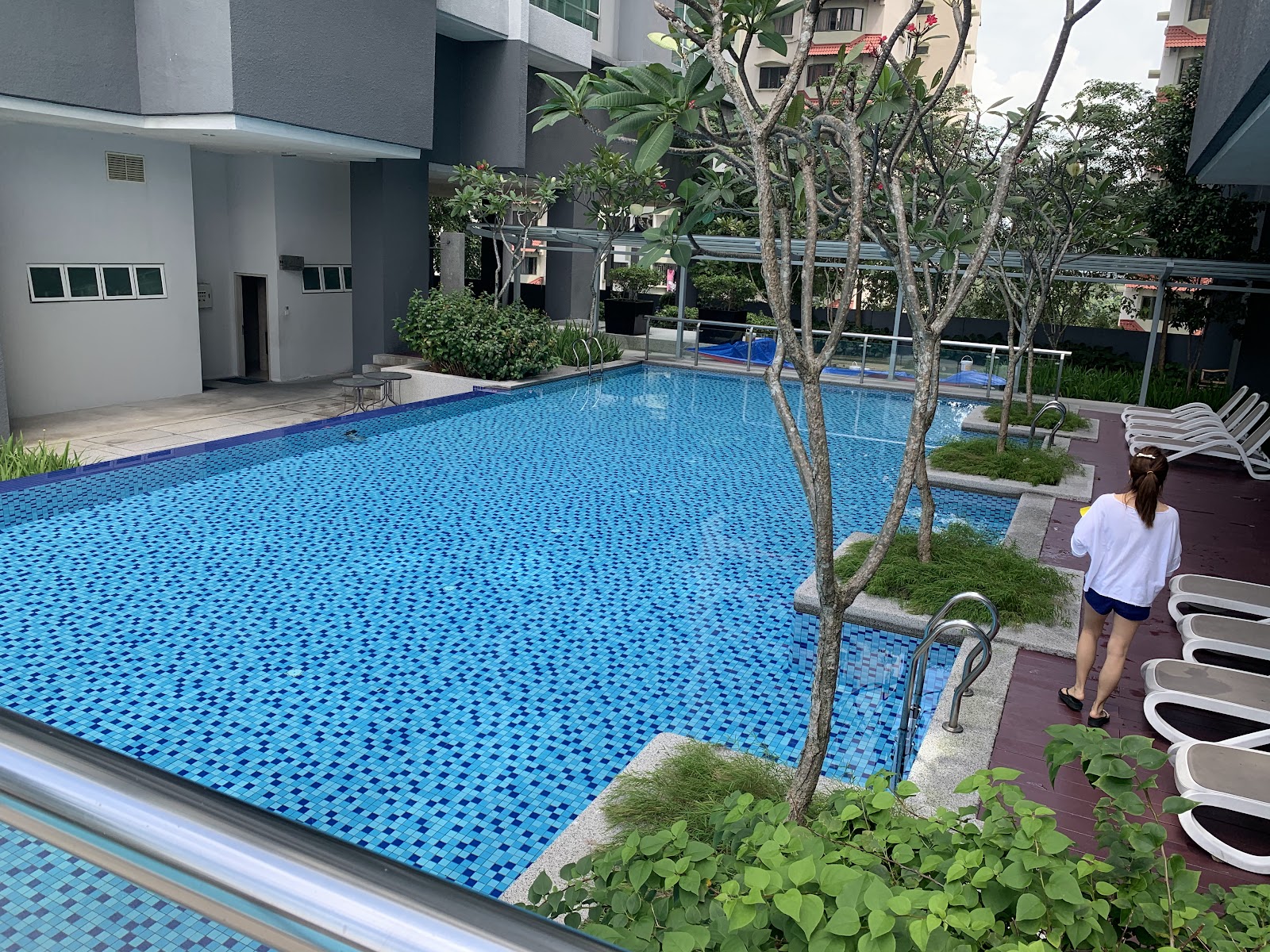 Glomac Damansara: A swimming pool area at a residential development with lounge chairs, greenery, and a person walking by
