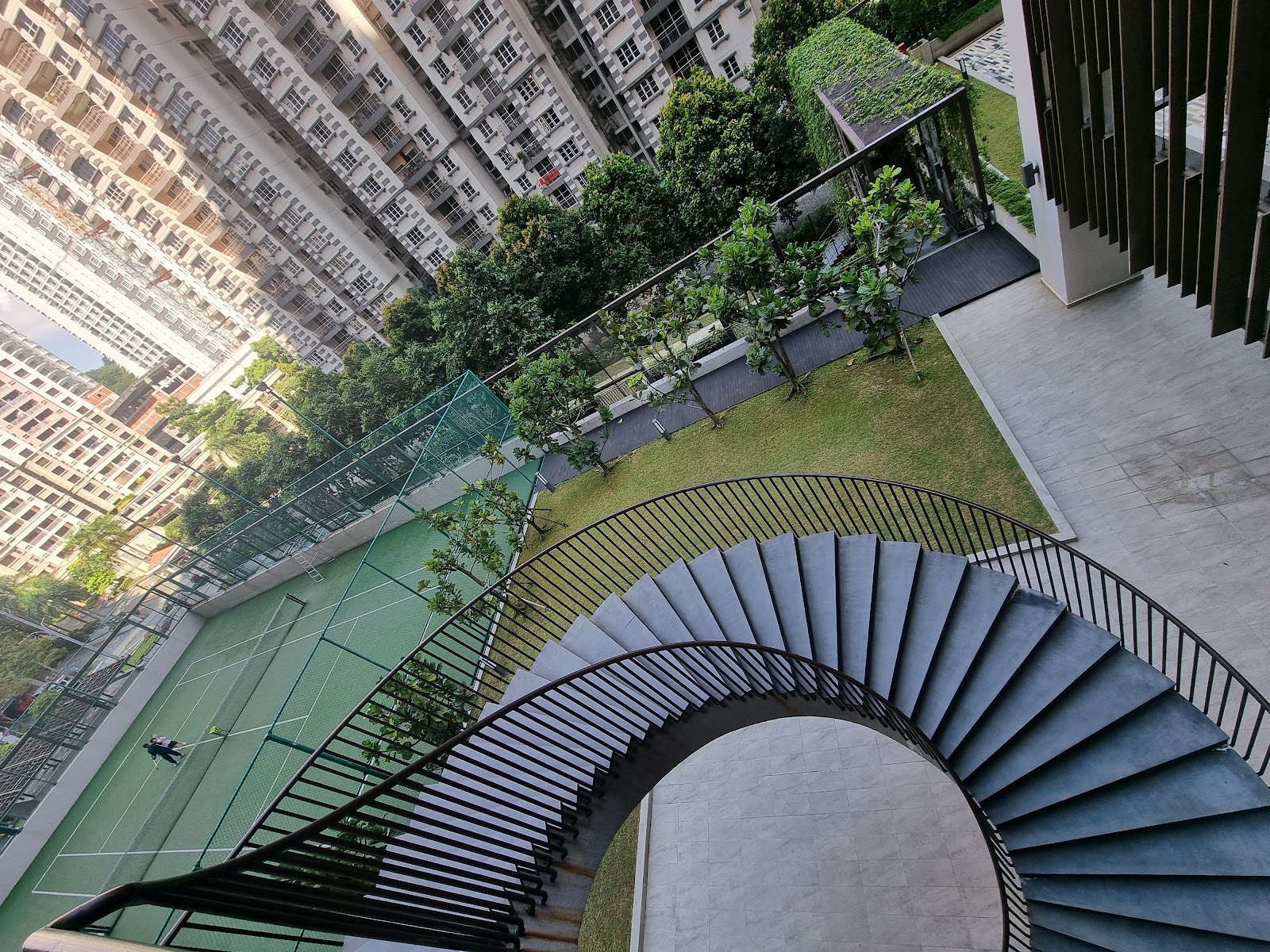 Residensi 22: An overhead view of a spiral staircase leading to a rooftop garden and tennis court, with surrounding high-rise buildings in the background