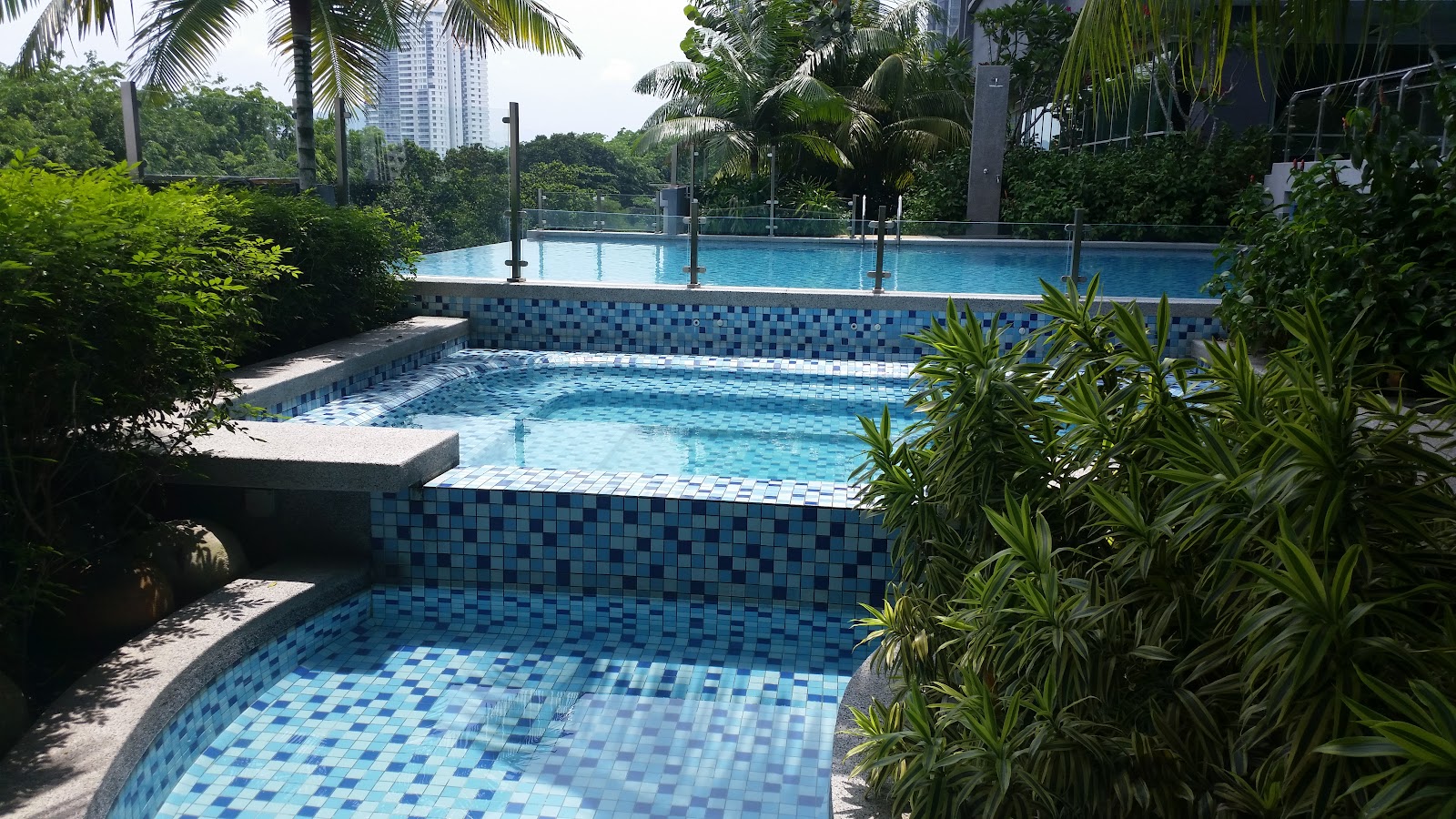 Glomac Damansara: Swimming pools with tiered tiled edges surrounded by tropical plants and trees, with a city high-rise visible in the background