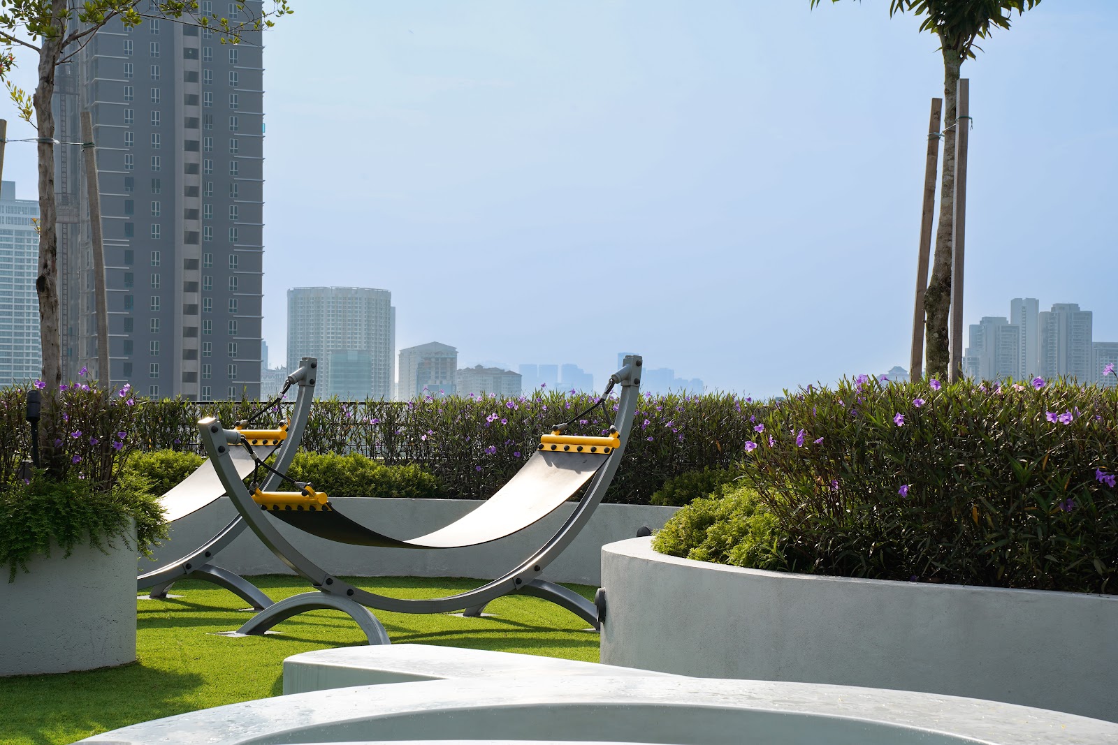 Utamara Boutique Residences: Rooftop garden area with hammock-style loungers, potted plants, and flowering shrubs, overlooking a city skyline with high-rise buildings
