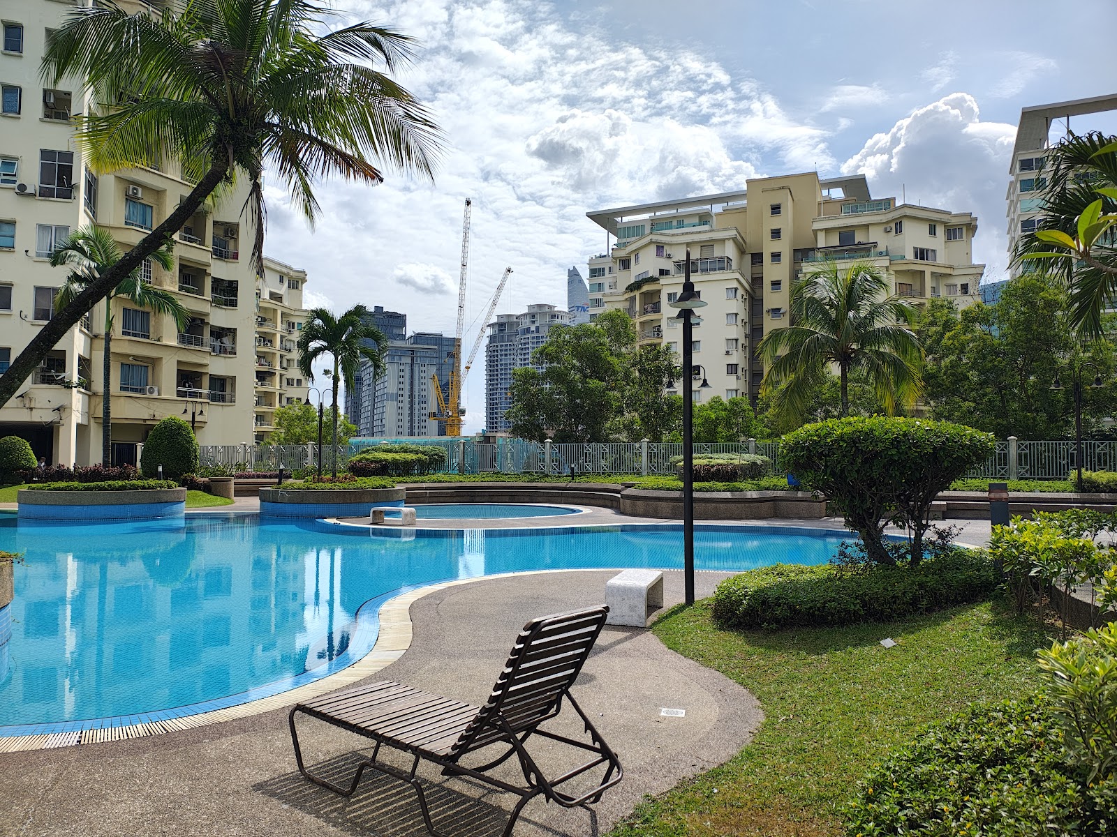 Pantai Panorama Condominiums: Swimming pool area of a condominium with loungers, palm trees, and city high-rises with a construction crane in the background