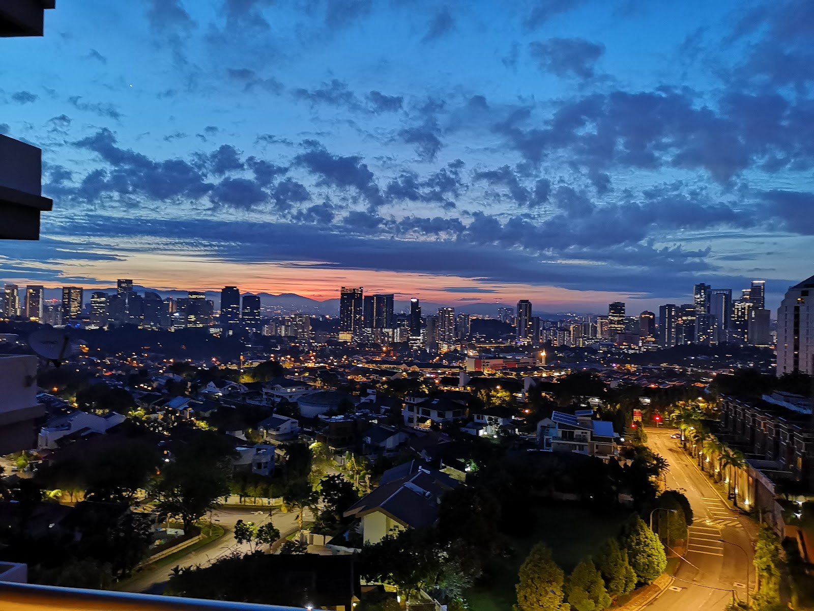 Park Rose: City skyline at dusk with sunset colors and illuminated buildings, viewed from a high-rise balcony overlooking suburban rooftops and streets below