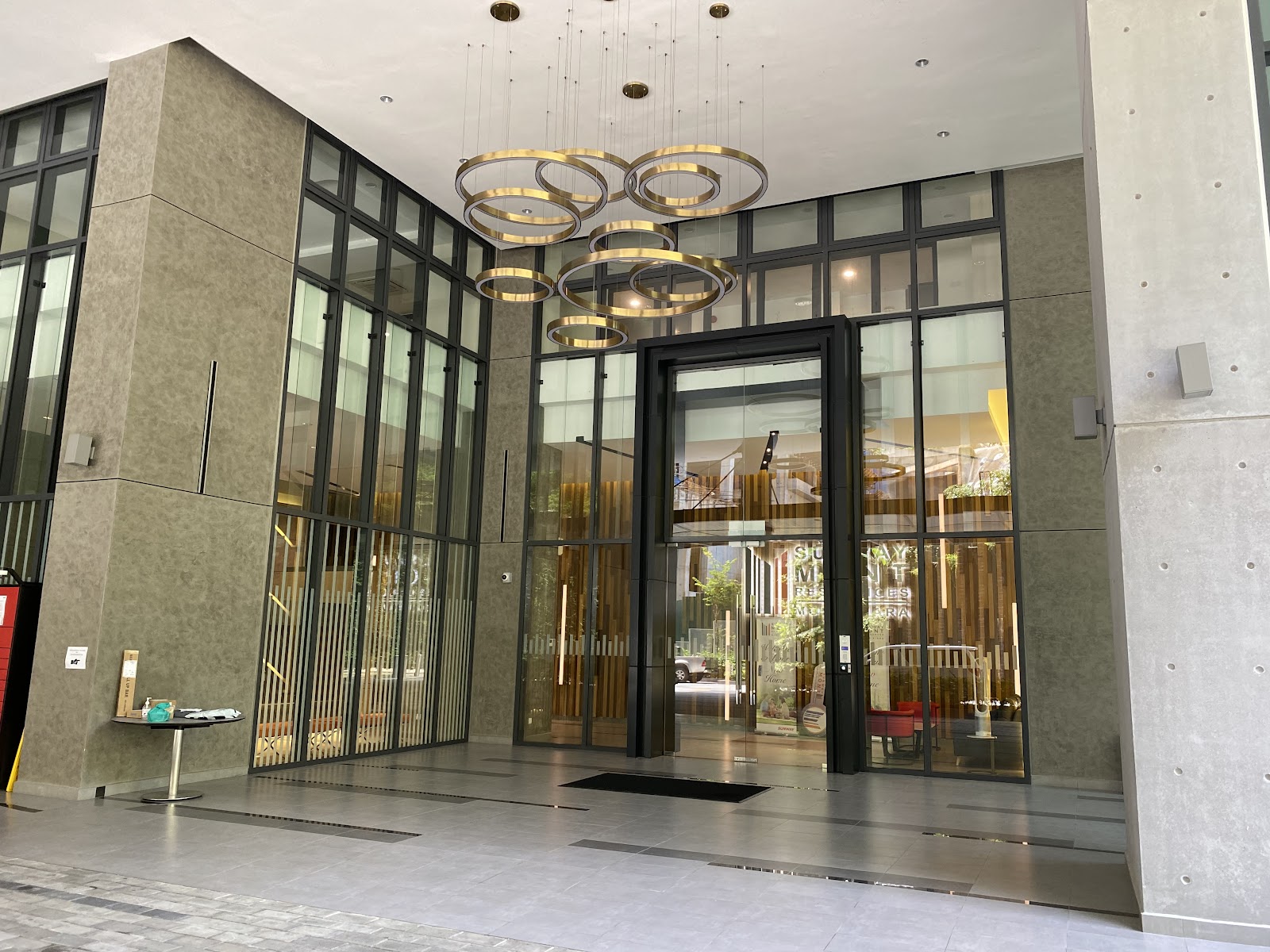Sunway Mont Residences: Entrance facade of a residential building with glass doors, gold ring pendant lights in the lobby ceiling, and a decorative wooden slat backdrop visible through the glass