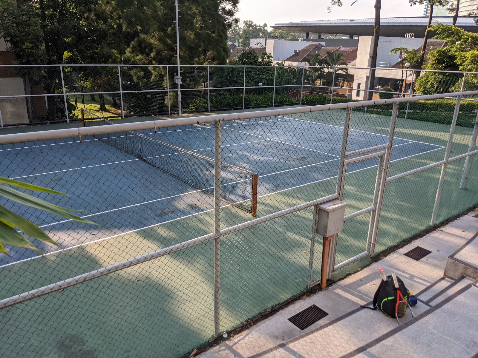 Bangsar Puteri: A tennis court surrounded by chain-link fencing, with a tennis bag and rackets on the pavement outside the court