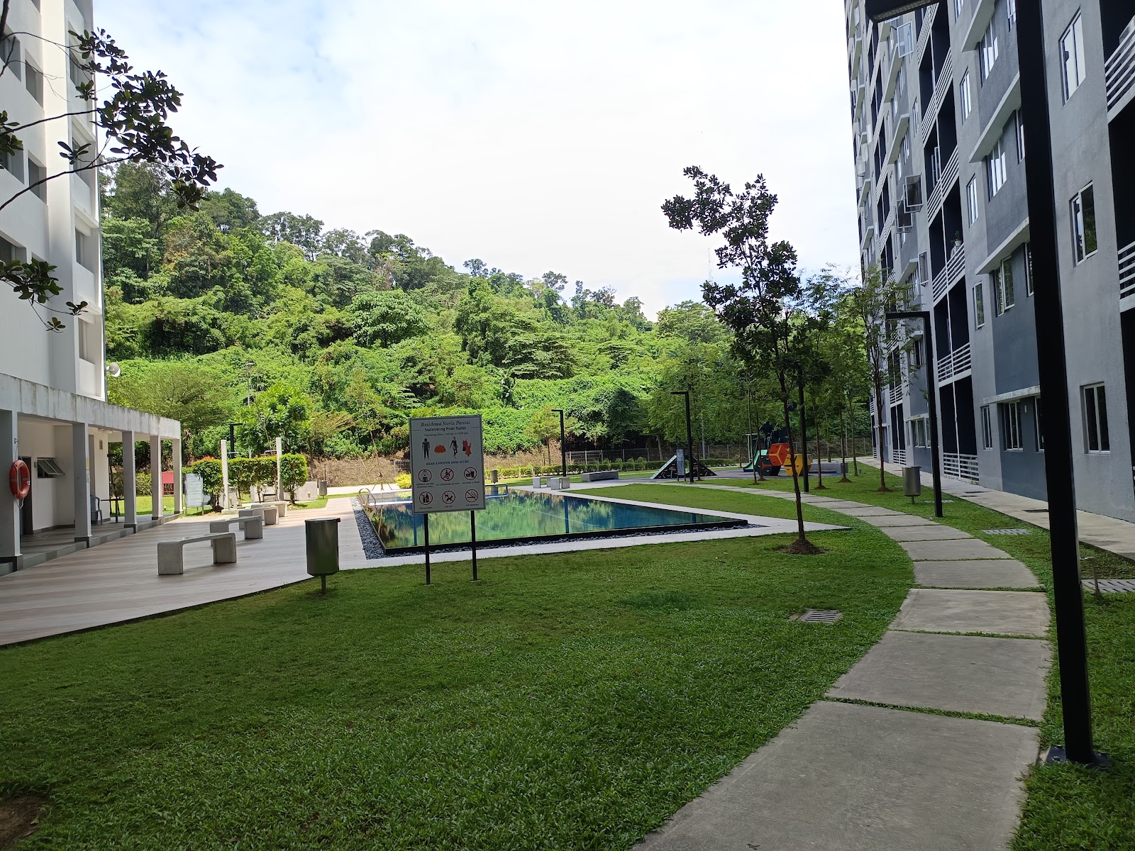 Suria Pantai Residency: Swimming pool and courtyard area between residential buildings with a sign, playground in the background, surrounded by greenery