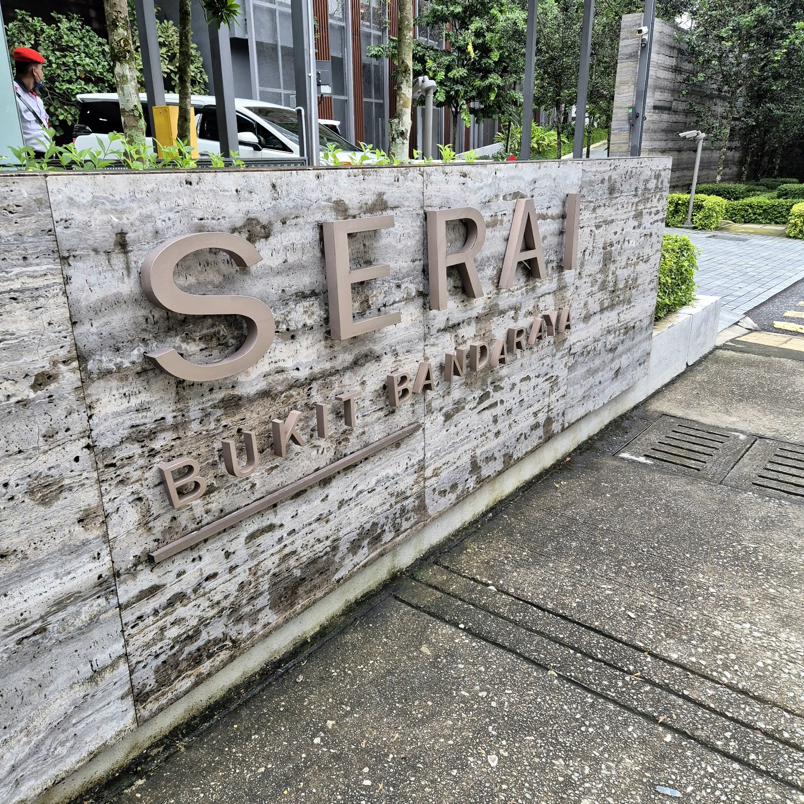 Serai: Entrance sign wall for 'Serai Bukit Bandaraya' development with a security guard and parked cars visible behind