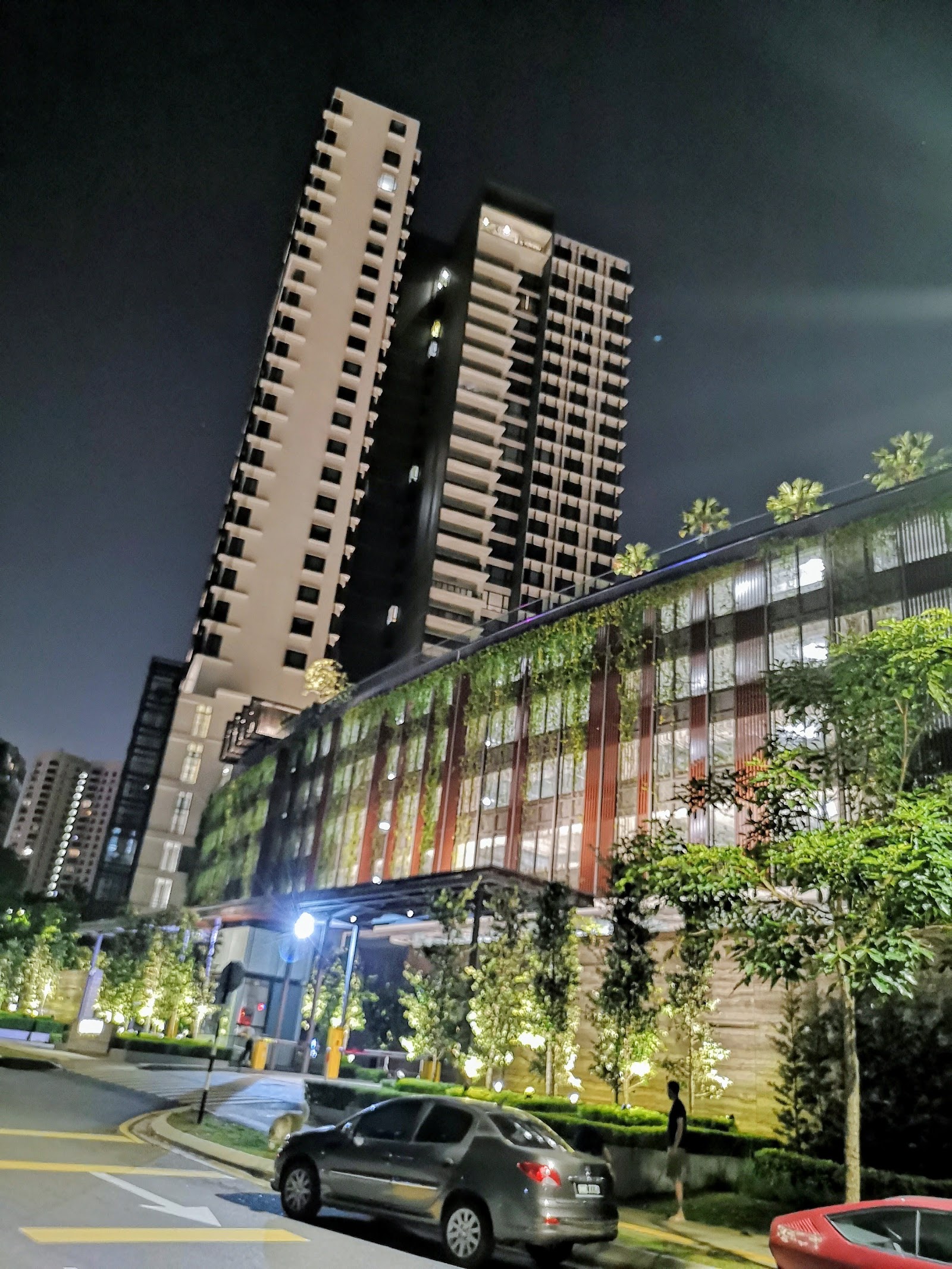 Serai: Nighttime view of a residential tower complex with a lush green vertical garden facade, cars parked on the street in front