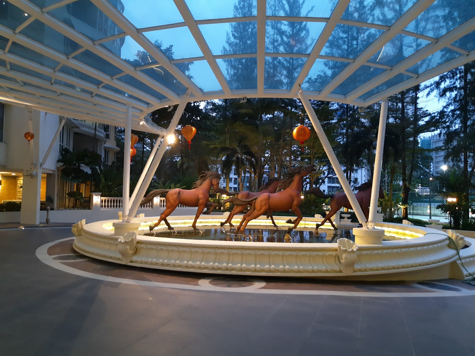 La Grande Kiara: A decorative fountain feature with bronze horse statues under a glass-and-steel canopy, surrounded by lanterns and greenery at dusk