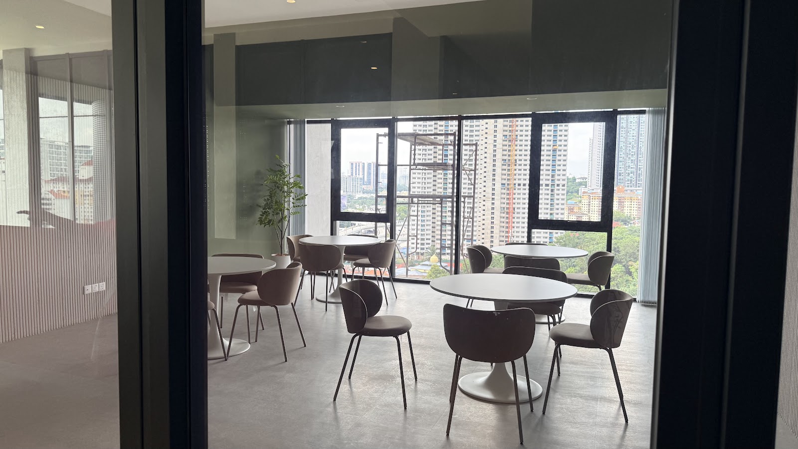 Nestree Residence: Communal dining/lounge area with round tables and chairs, viewed through glass doors, with a city skyline visible through floor-to-ceiling windows