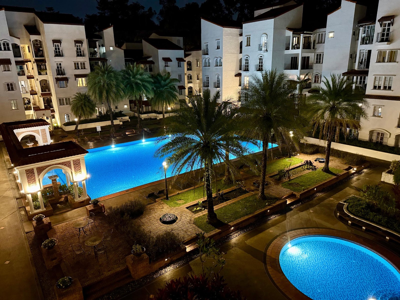 Andalucia: Nighttime view of a resort-style swimming pool with palm trees, lounge chairs, and a small circular pool in the foreground, surrounded by residential apartment buildings