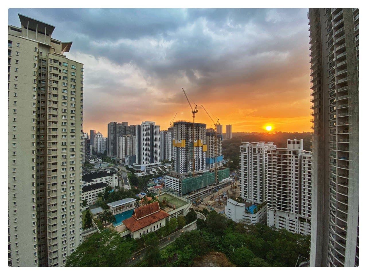 28 Mont Kiara: Sunset skyline view from a high-rise balcony showing surrounding towers, construction cranes, and a low-rise building with a red-tiled roof and pool below