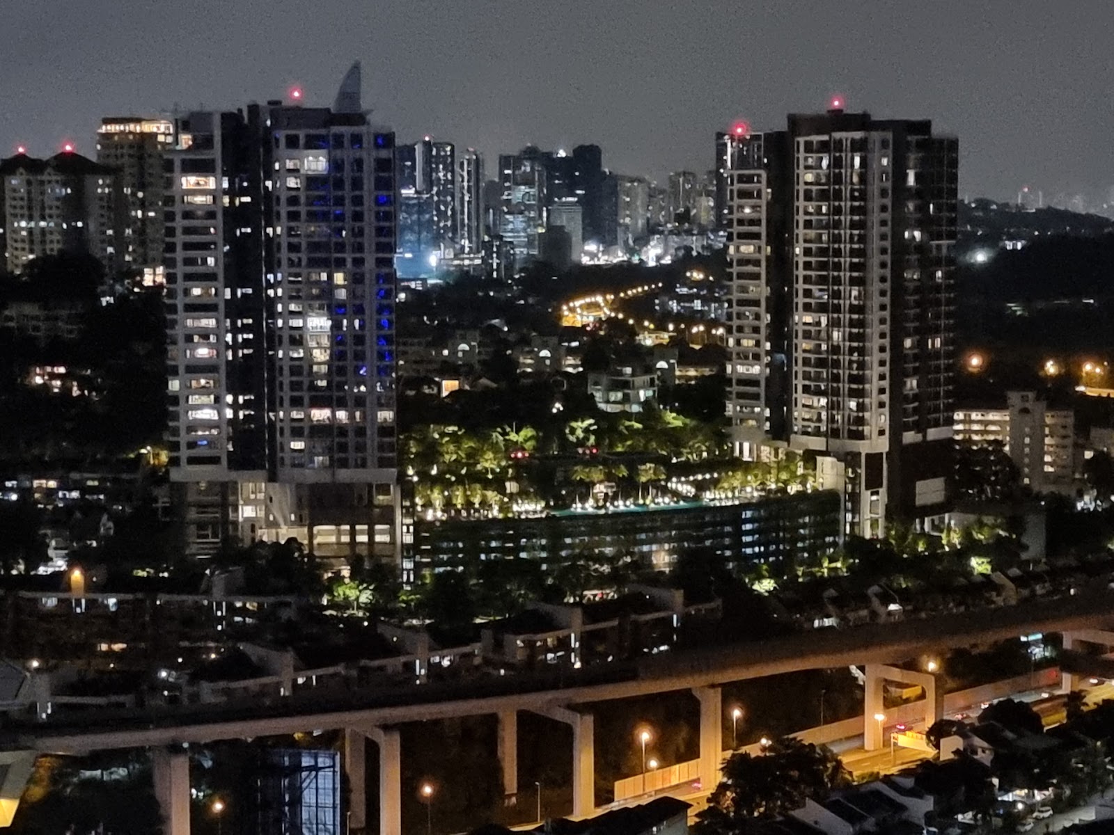 Serai: Night-time cityscape view of illuminated high-rise towers, a pool and gardens, with an elevated highway in the foreground