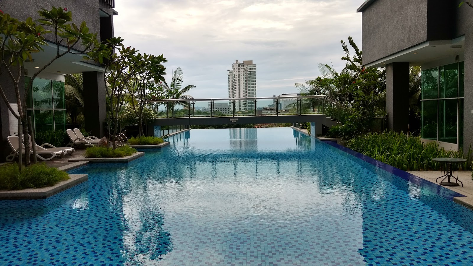 Glomac Damansara: An infinity-edge swimming pool at a residential development, with a bridge walkway, loungers, and a city skyline in the background