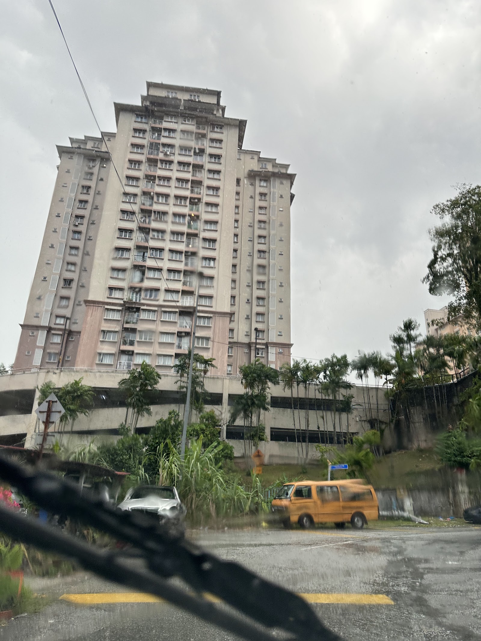 Sri TTDI: A tall residential tower viewed from a rainy street, with palm trees, a parking structure base, and vehicles in the foreground