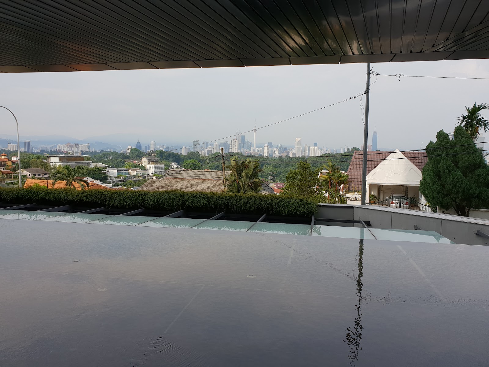 Rhombus @ Bangsar: Skyline view of Kuala Lumpur city with towers and greenery, seen from a rooftop pool deck with hedges and a metal shade structure overhead