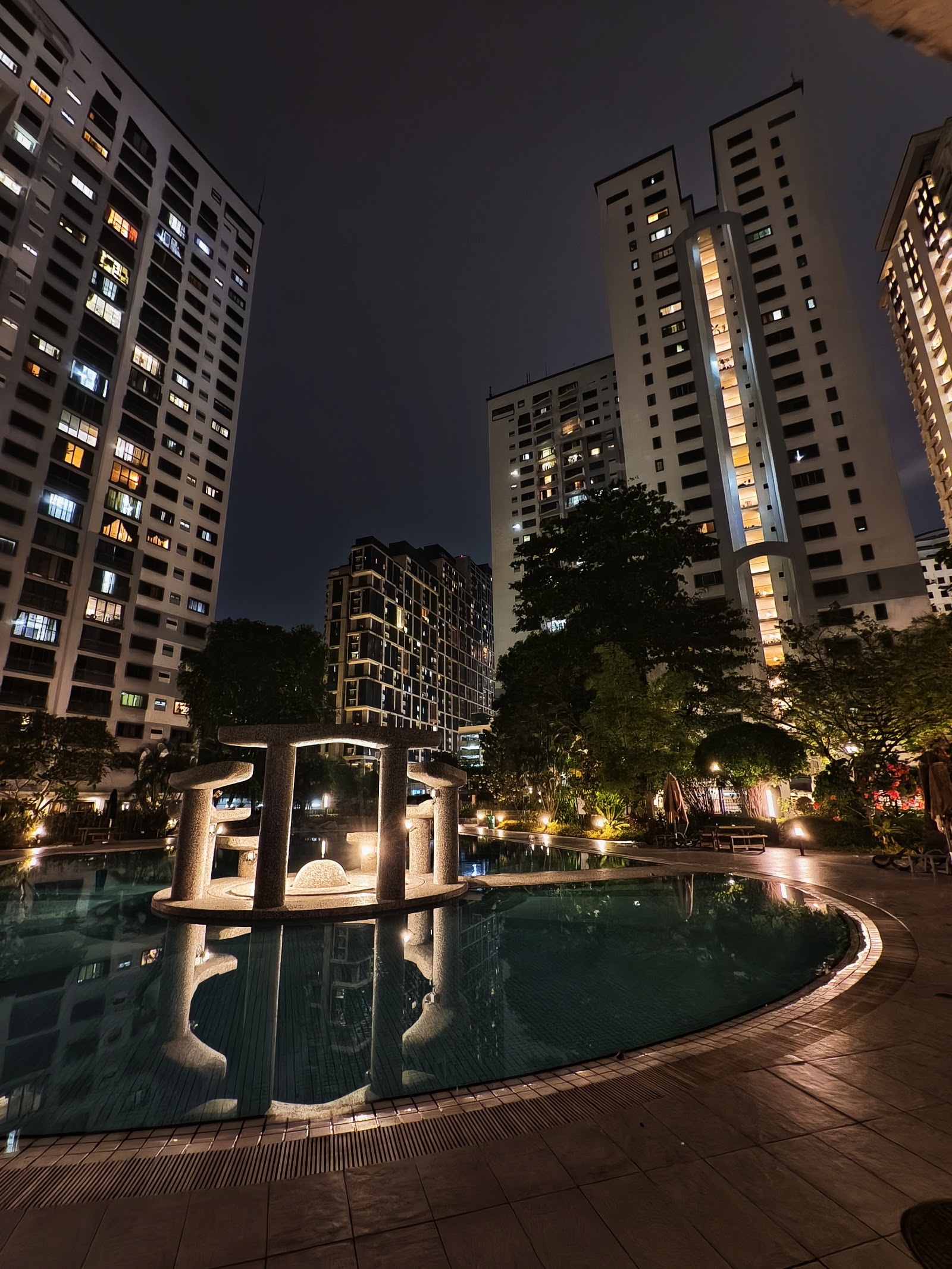 Vista Kiara: Nighttime view of a swimming pool with a stone gazebo structure in the middle, surrounded by illuminated high-rise residential towers