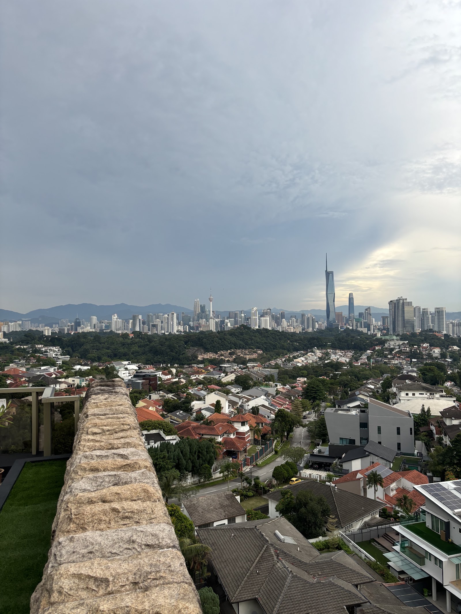 The Loft: City skyline view with skyscrapers, including the Petronas Twin Towers-area cluster and KL Tower, seen from a rooftop with a stone parapet overlooking residential rooftops and greenery