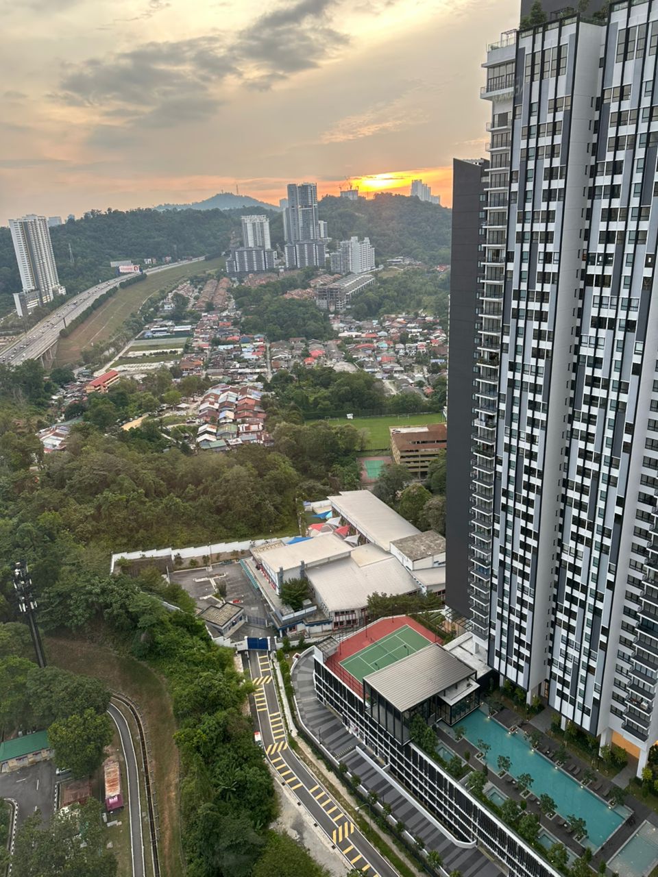 Residensi Dutamas Dahlia: Aerial view from a high-rise balcony showing the pool, tennis court and facility deck below, with surrounding neighborhood, highway, and hills under a sunset sky