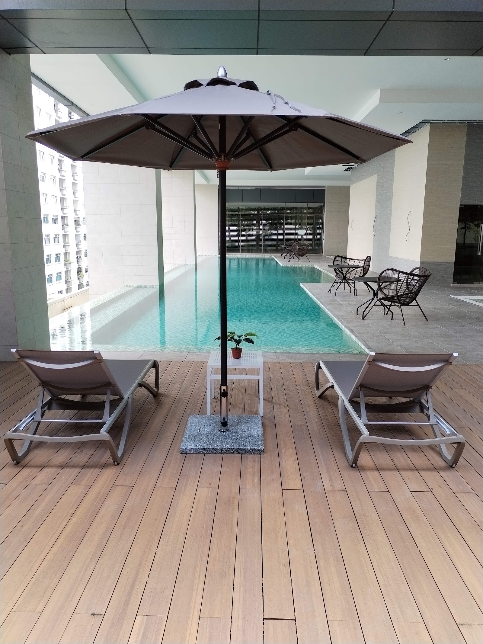 Pinnacle Bangsar: Poolside deck with lounge chairs, umbrella, and dining chairs beside an indoor infinity pool overlooking city buildings