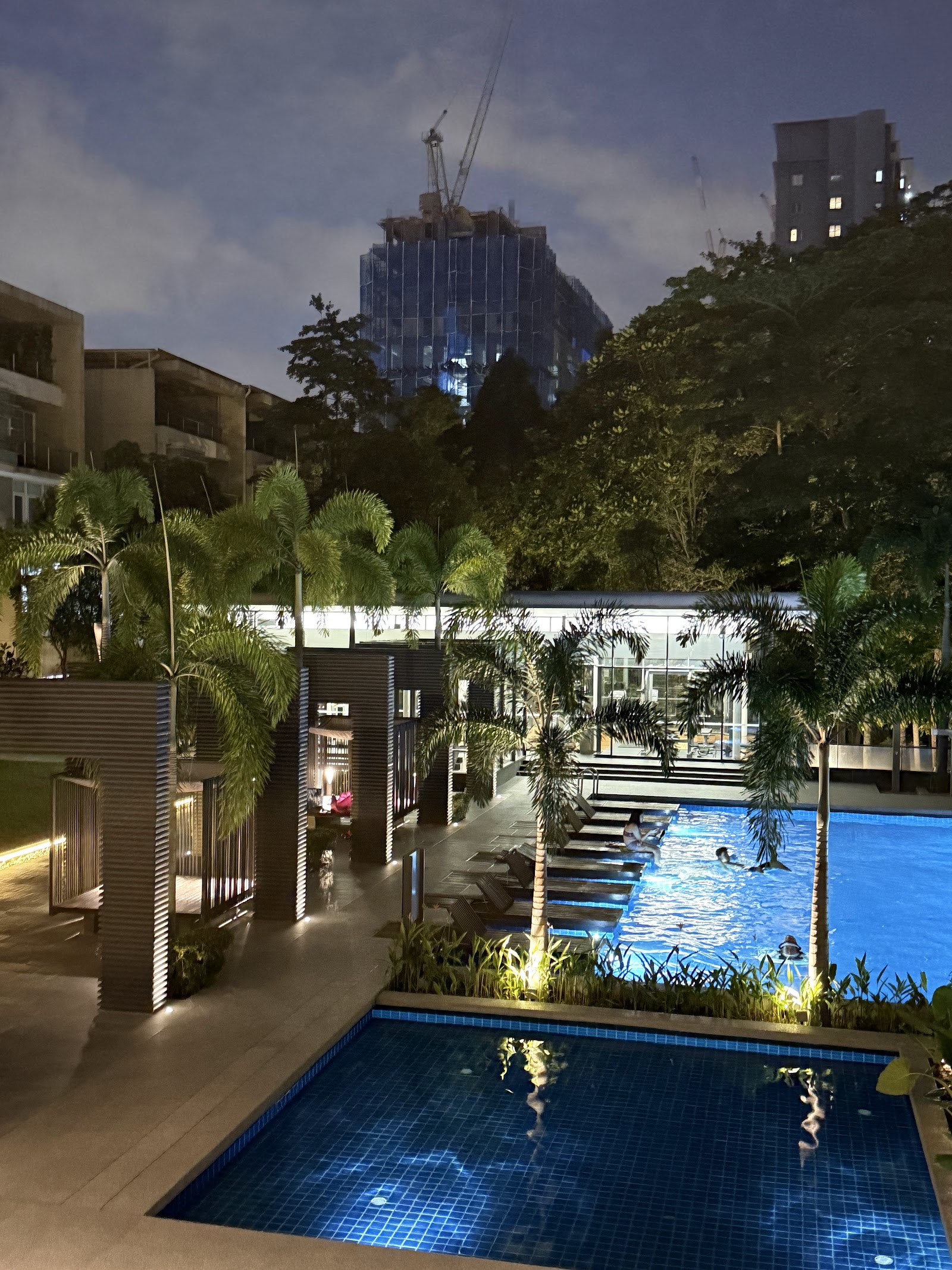One Menerung: Nighttime view of a swimming pool area with lounge chairs, palm trees, and a poolside pavilion, with city buildings in the background