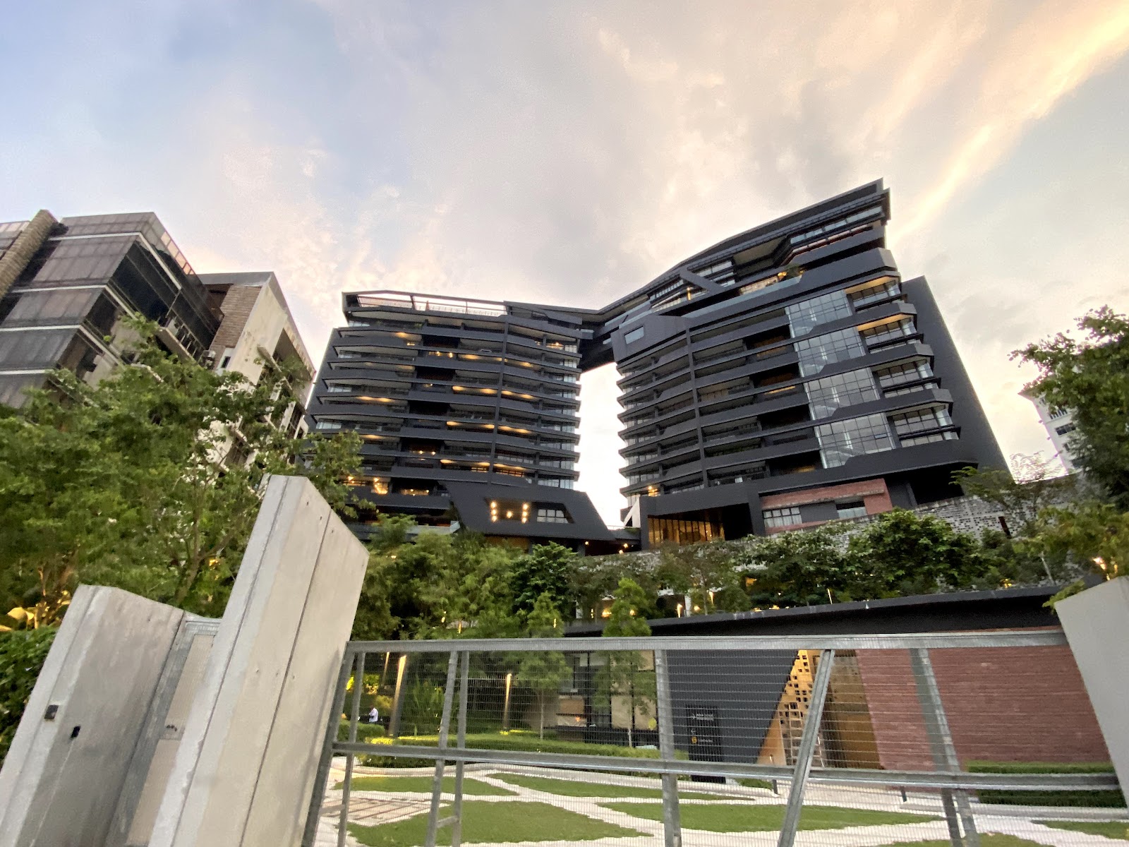Kapas Heights: Modern high-rise residential towers with dark facade and cantilevered upper floors, viewed from behind a fence with landscaped grounds in foreground